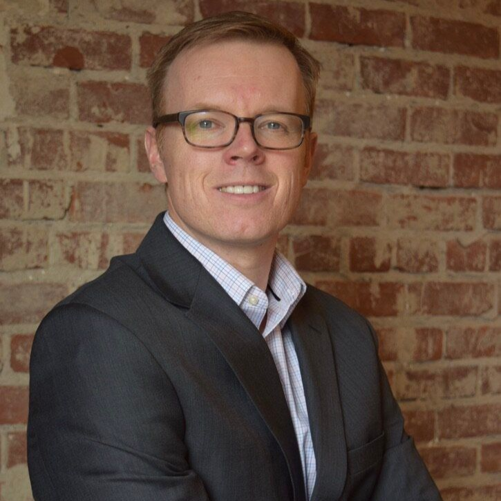 Man with glasses in suit jacket smiles in front of a brick wall.
