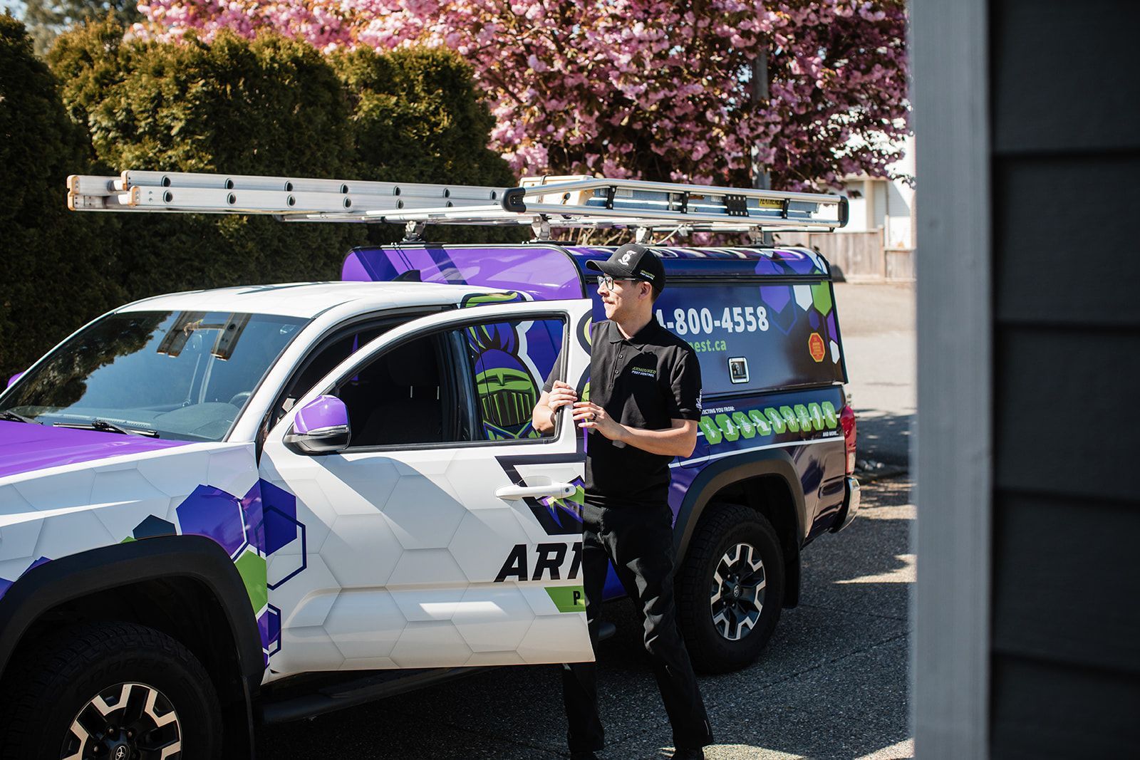 Person standing beside a white-and-purple utility truck with a ladder on top, in a sunny parking lot
