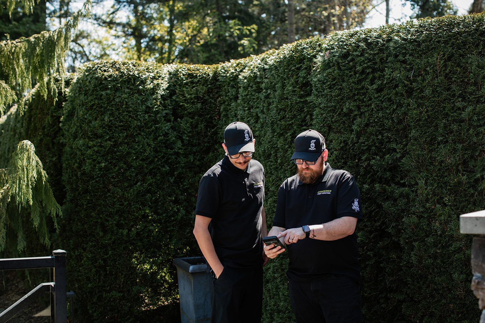 Two people in black clothing looking at a phone in front of a tall hedge outdoors