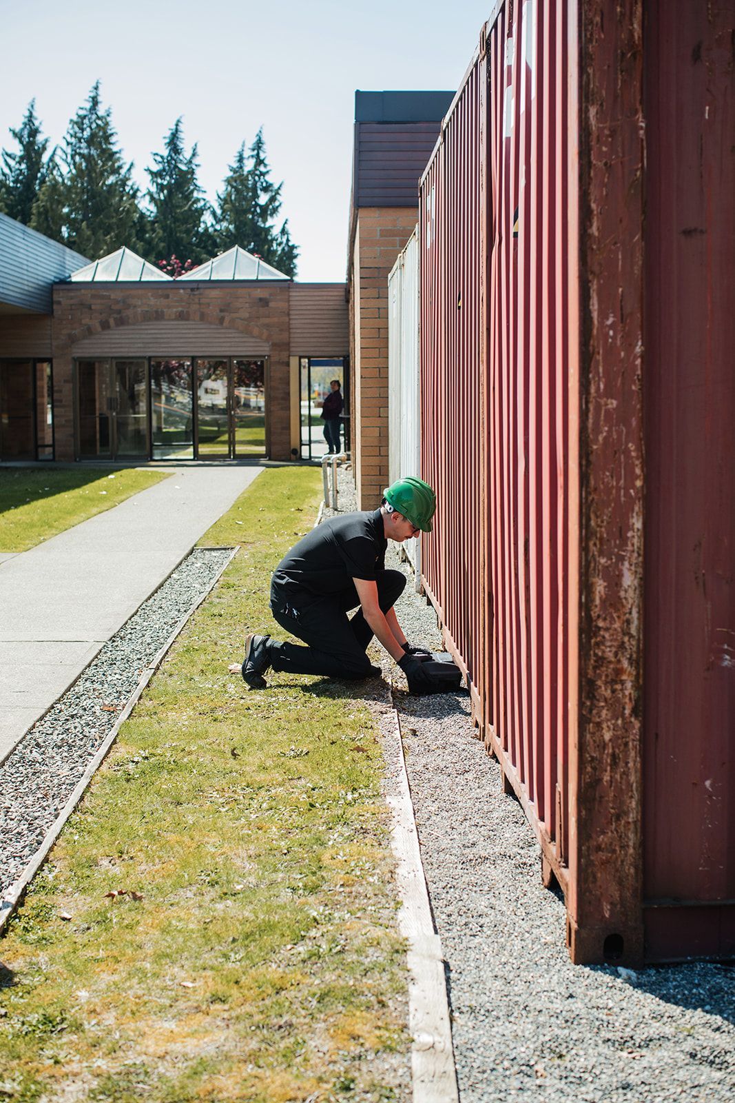 Person kneeling by a red wall, working along a gravel edge beside a building walkway.