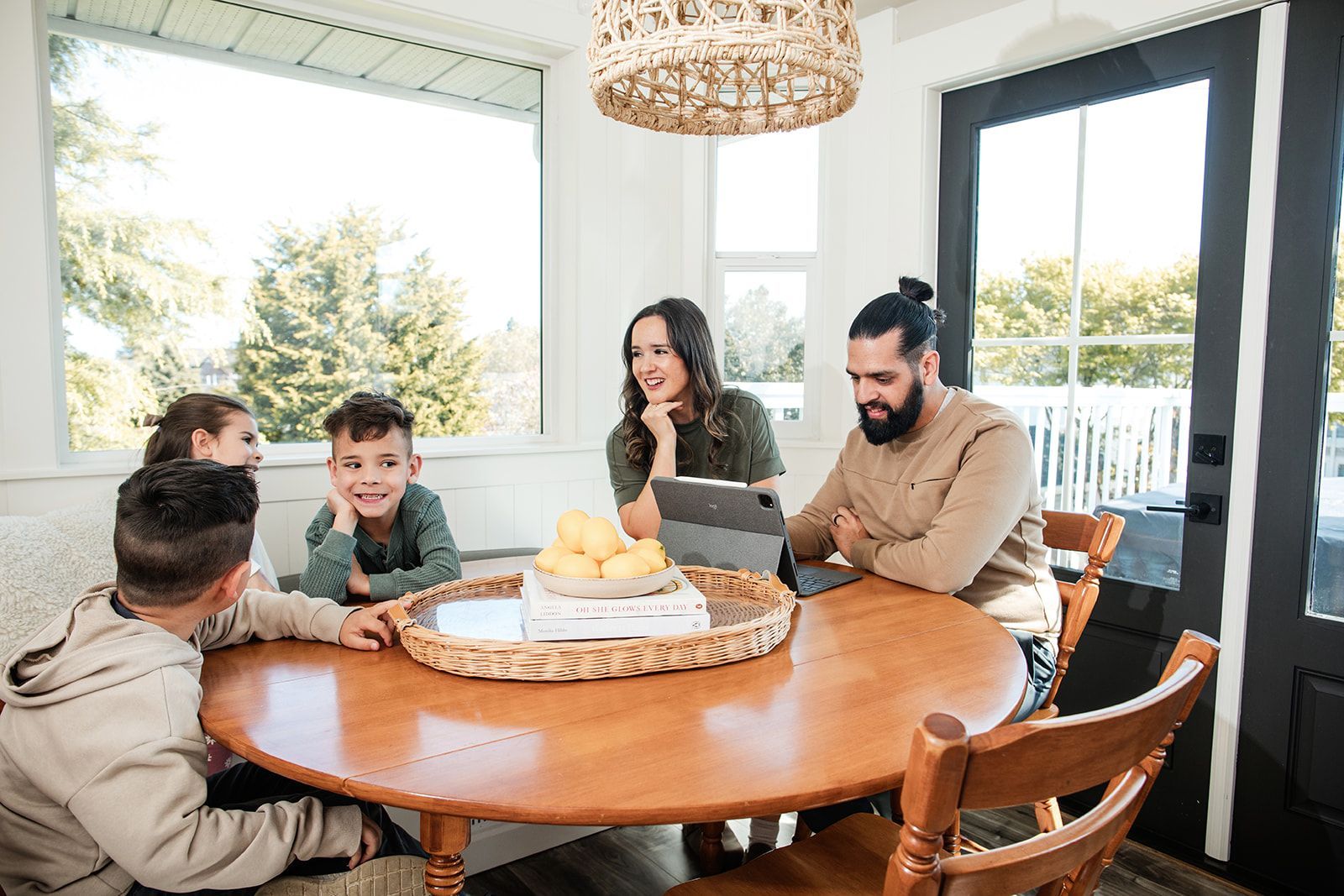 Family gathered around a wooden dining table in a bright sunroom, smiling and chatting over a cake.