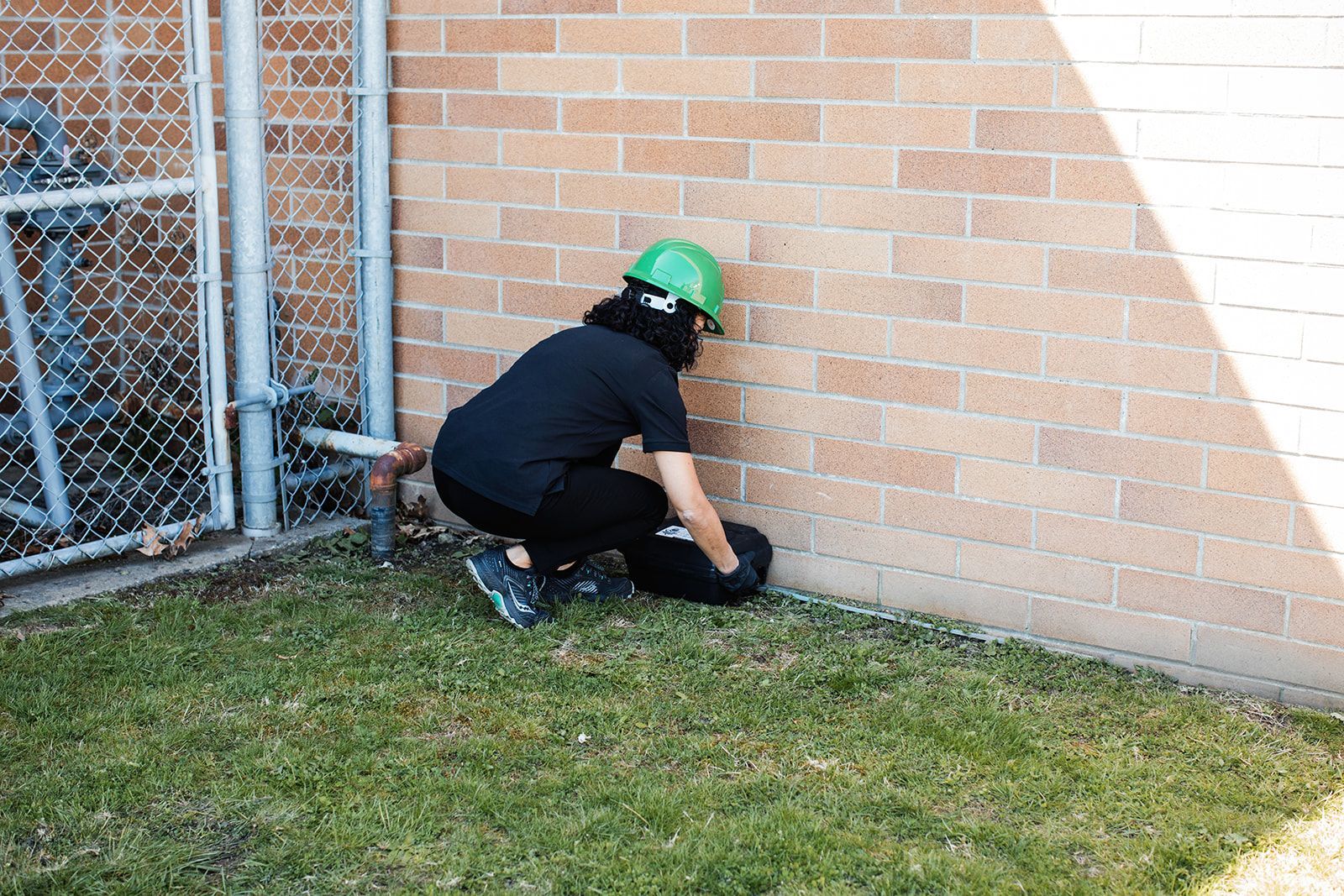 Worker in green hard hat kneeling by a brick wall, inspecting the ground near a fence and grass.