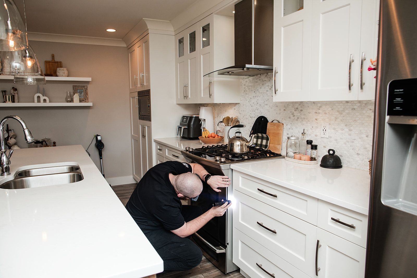 Person crouching in a bright white kitchen, inspecting an oven under the counter