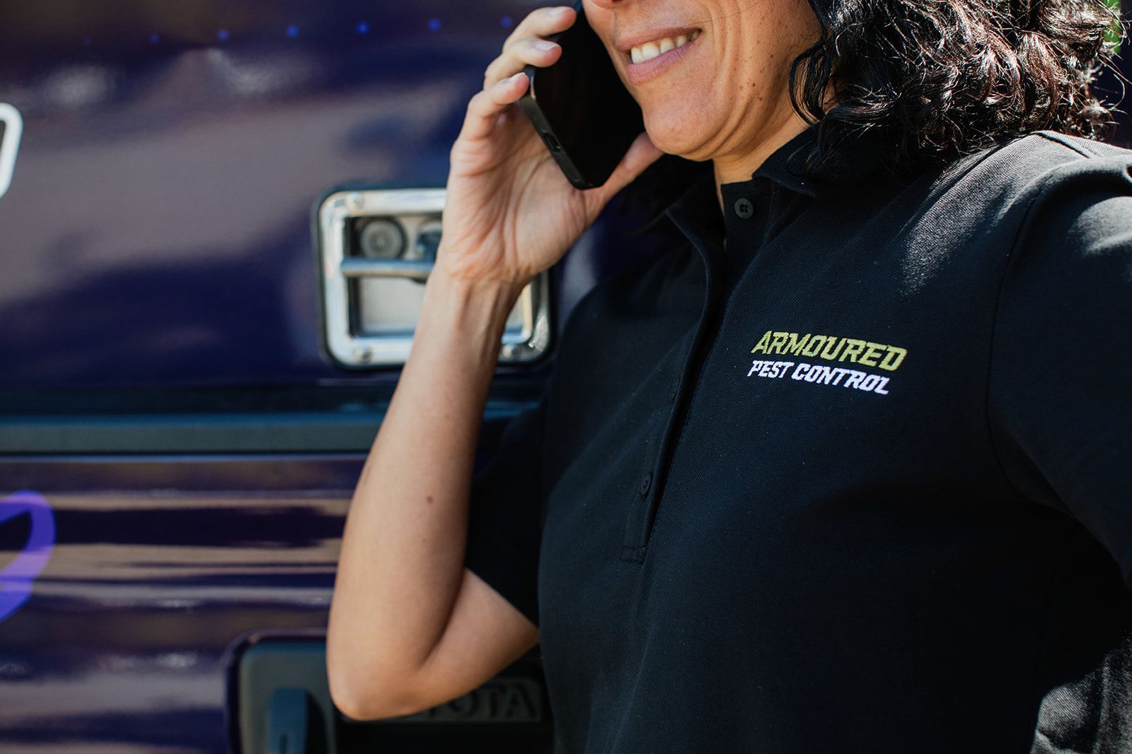 Woman in black shirt with company logo on phone, standing next to a vehicle.