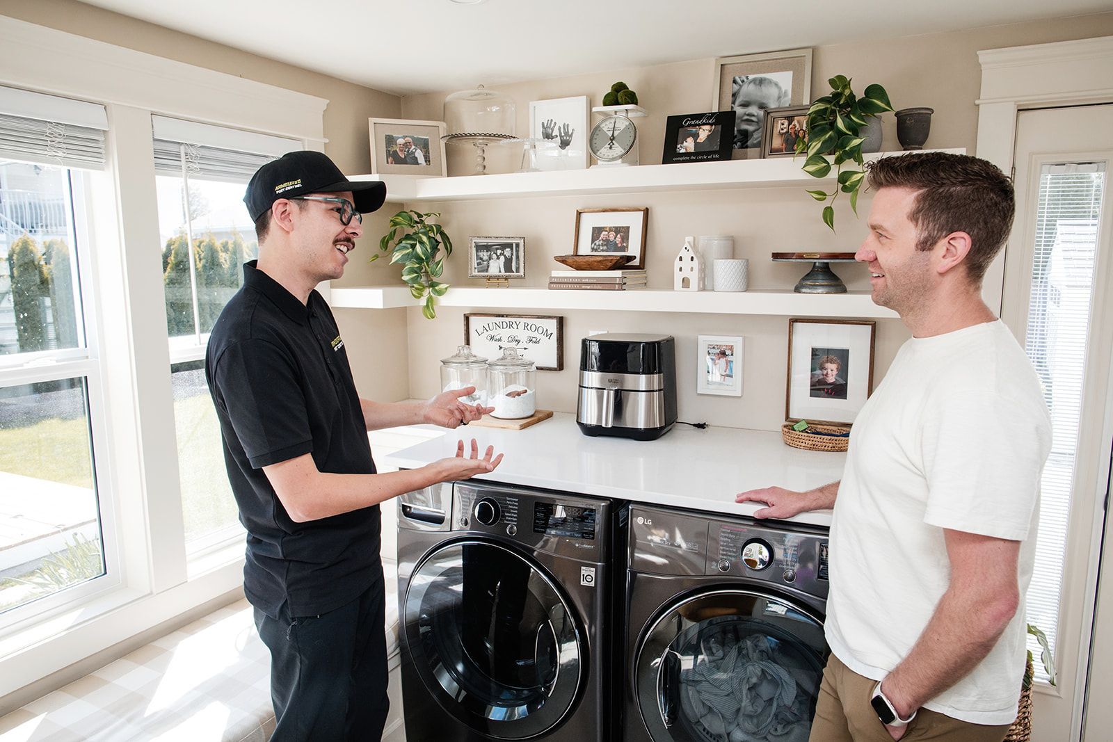 Two men talking in a laundry room beside black washer and dryer under white shelves