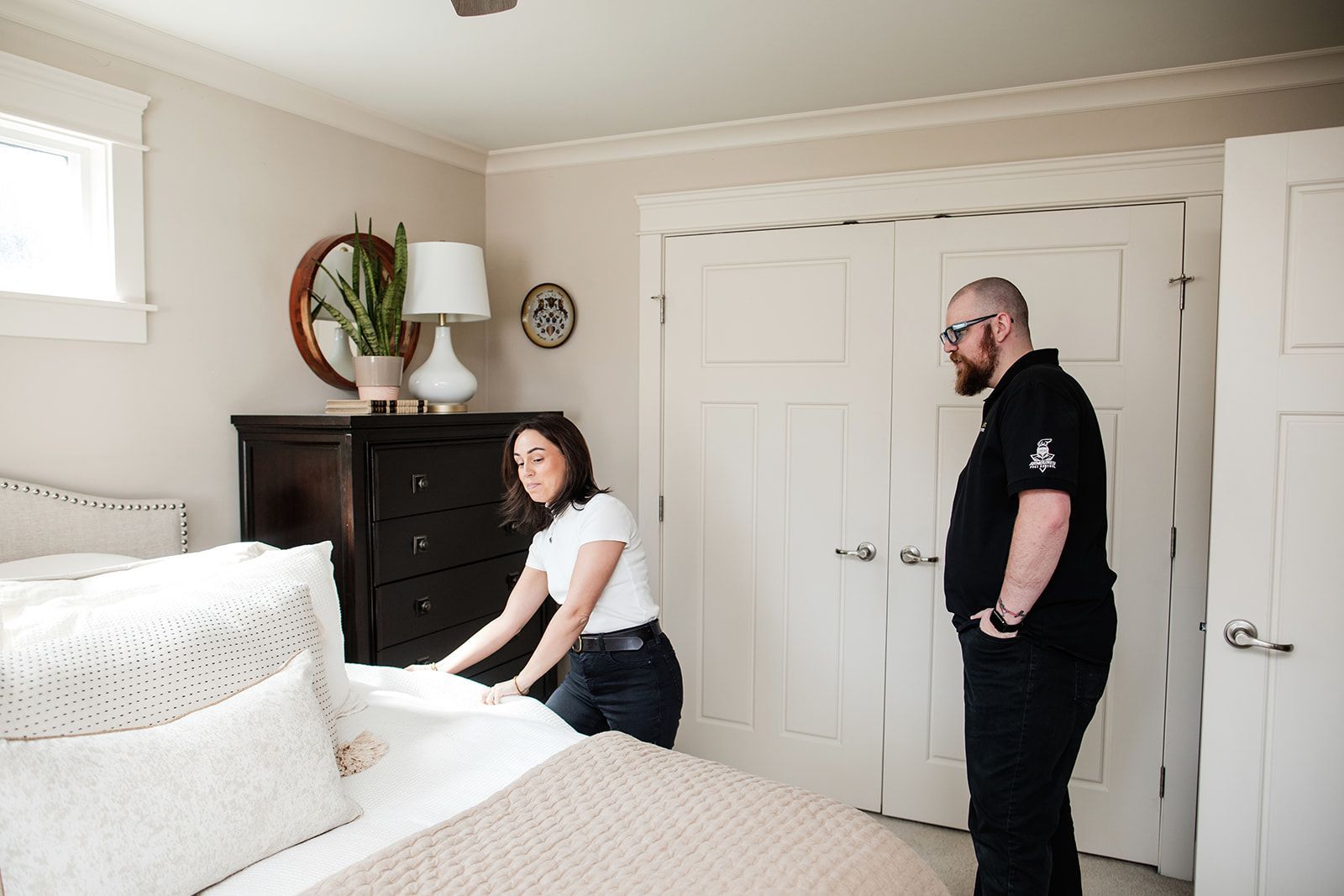 Two people in a bright bedroom, one adjusting a white bedspread near a black dresser and double closet doors.