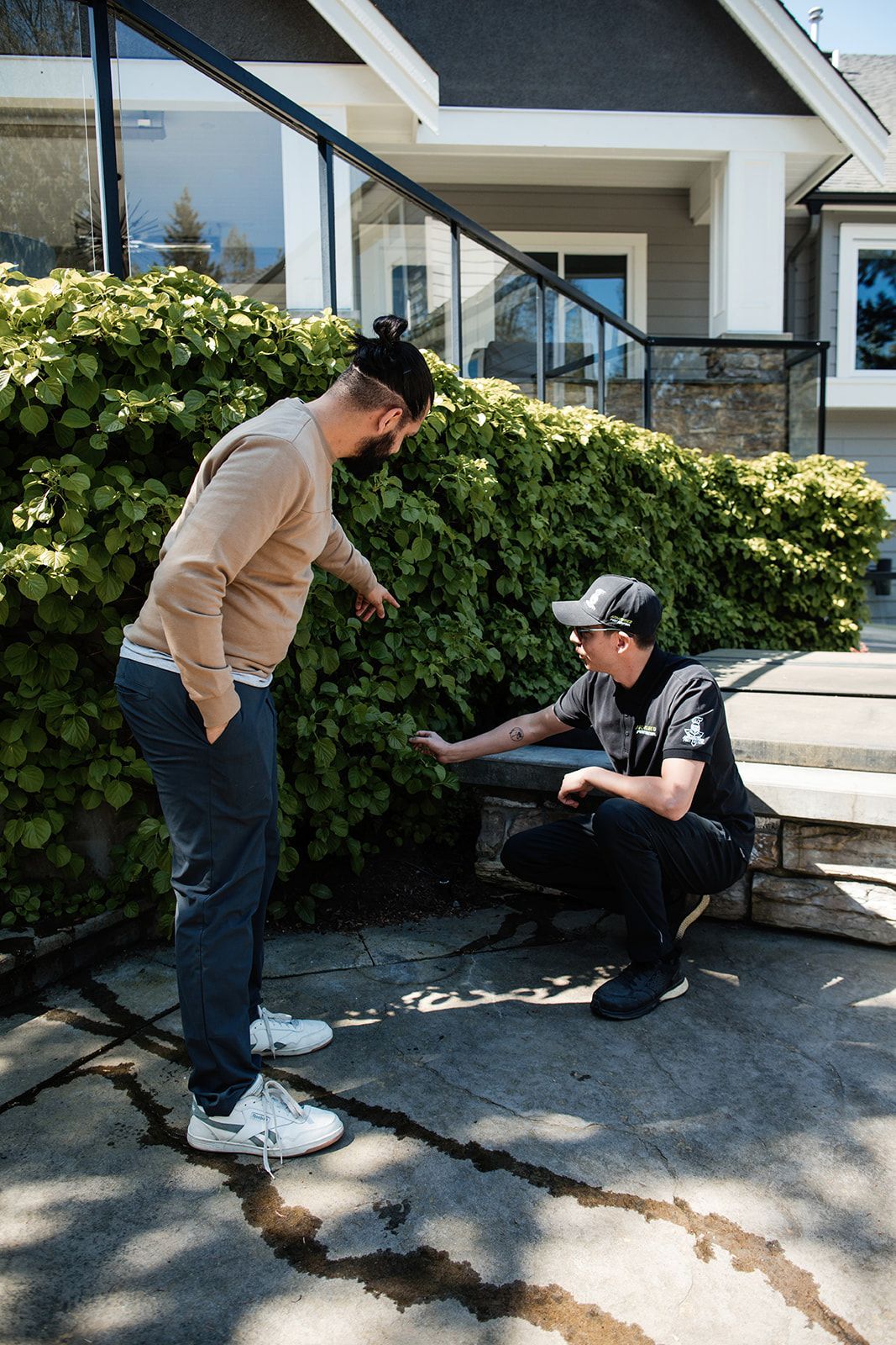Two people talking outside a house, one standing and one sitting on the curb near bushes.