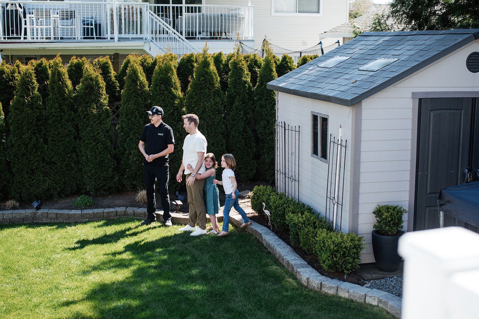 Three people and a child standing on a lawn beside a white garage and hedge-lined yard