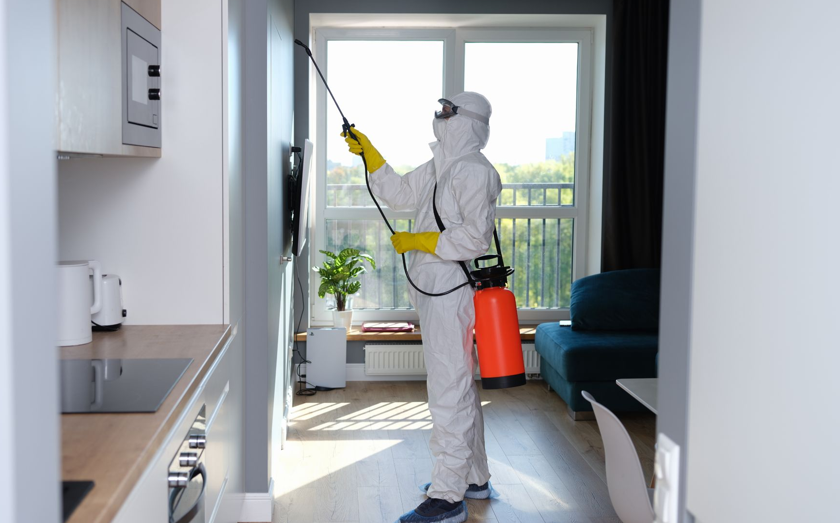 Worker in white protective gear spraying a balcony inside an apartment, holding a red tank sprayer.