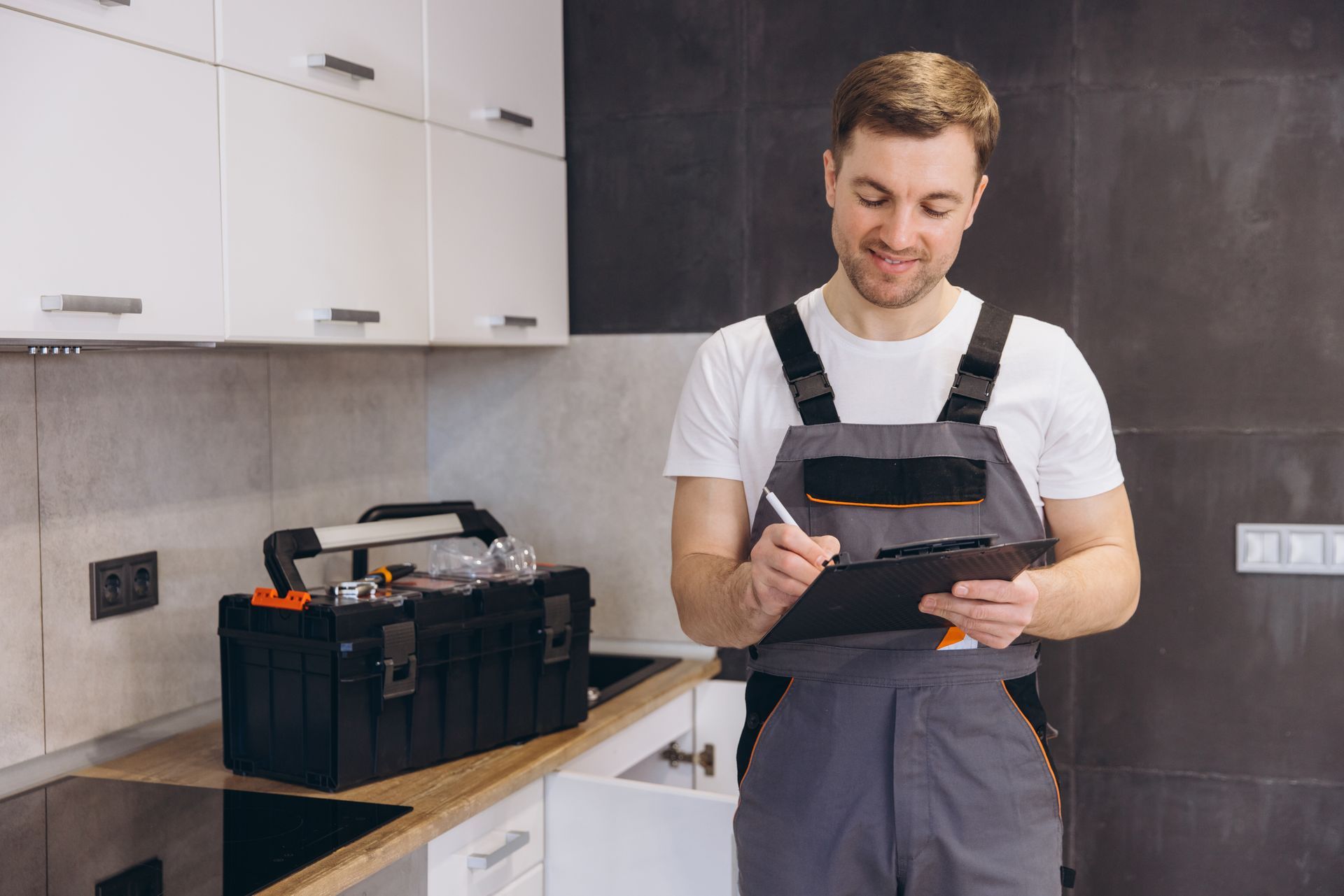 Technician in gray overalls checks a tablet beside a tool bag in a modern kitchen.