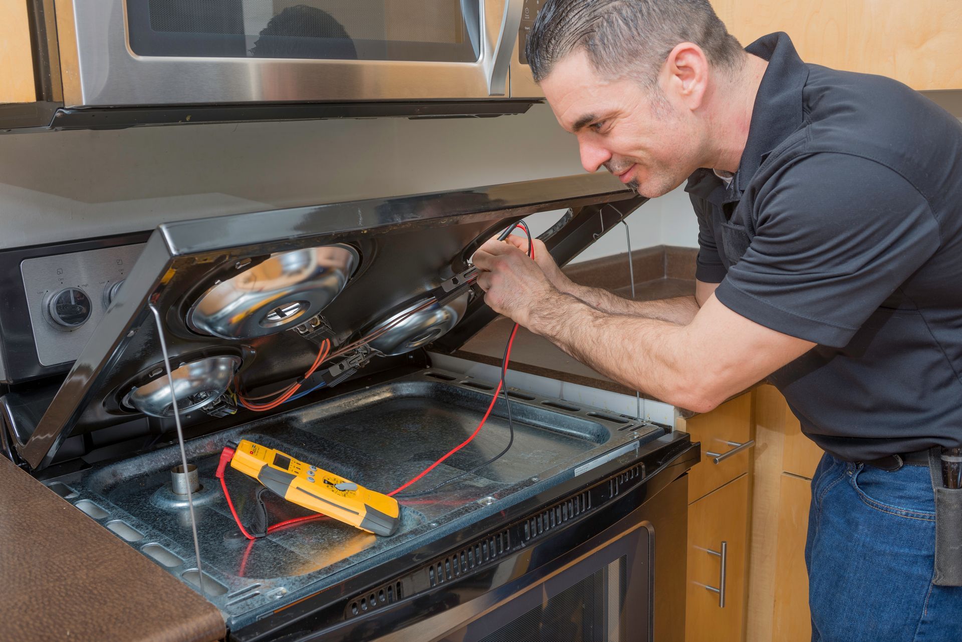 Man repairing a broken oven with tools, inspecting exposed wiring in a kitchen.