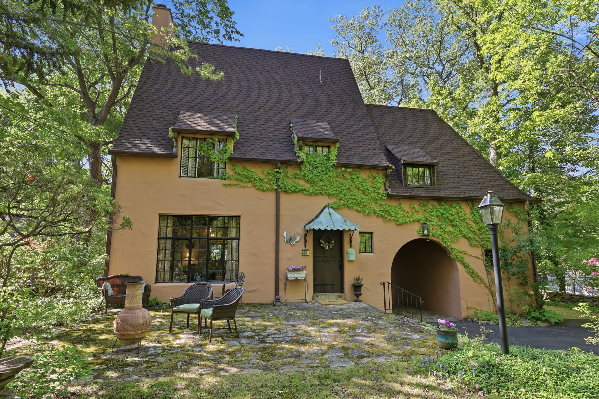Cottage with thatched roof, ivy-covered walls, and patio chairs in a sunny wooded garden