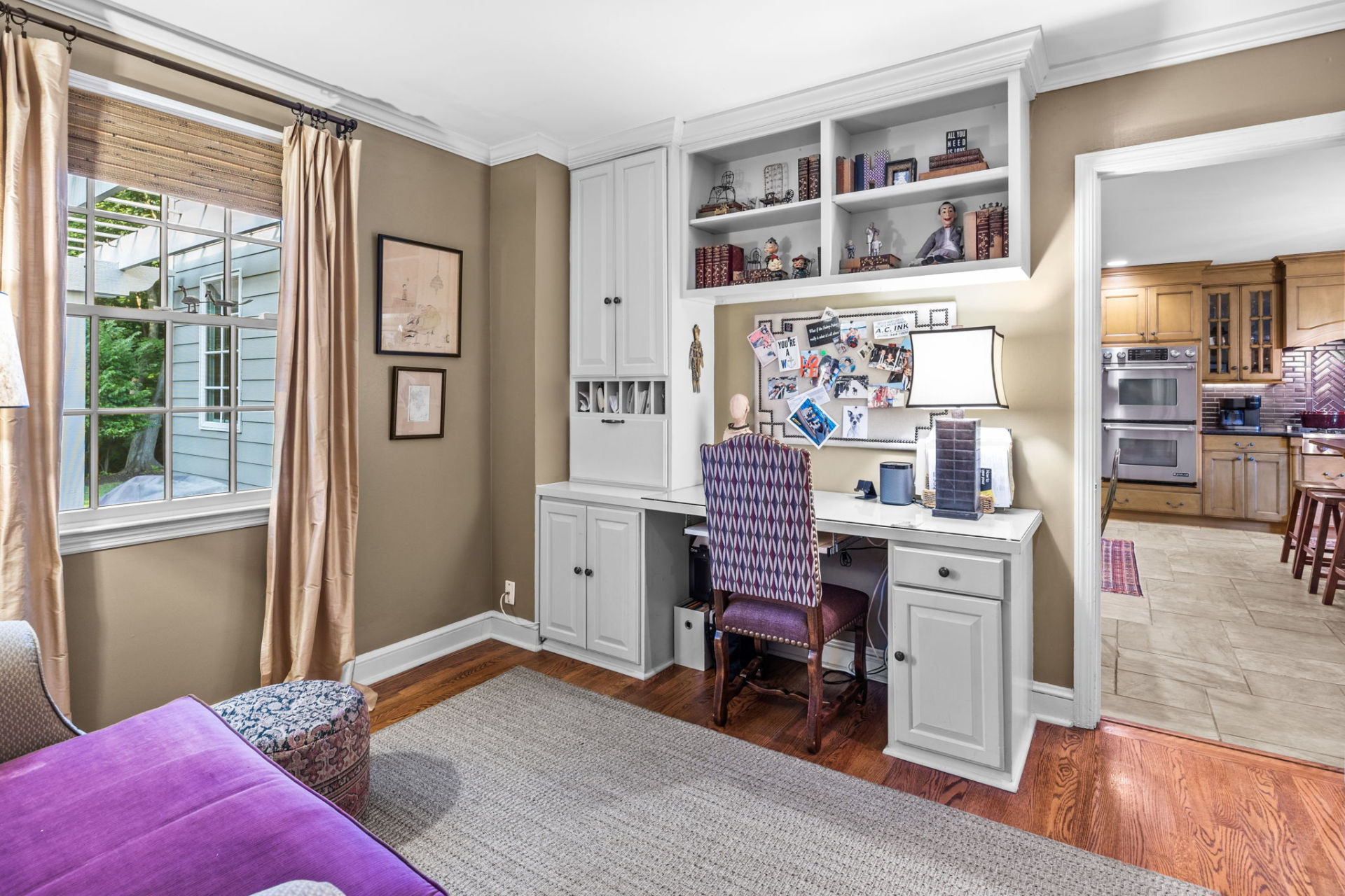 Home office with built-in desk, shelving, and window; purple chair; doorway to kitchen with oven.