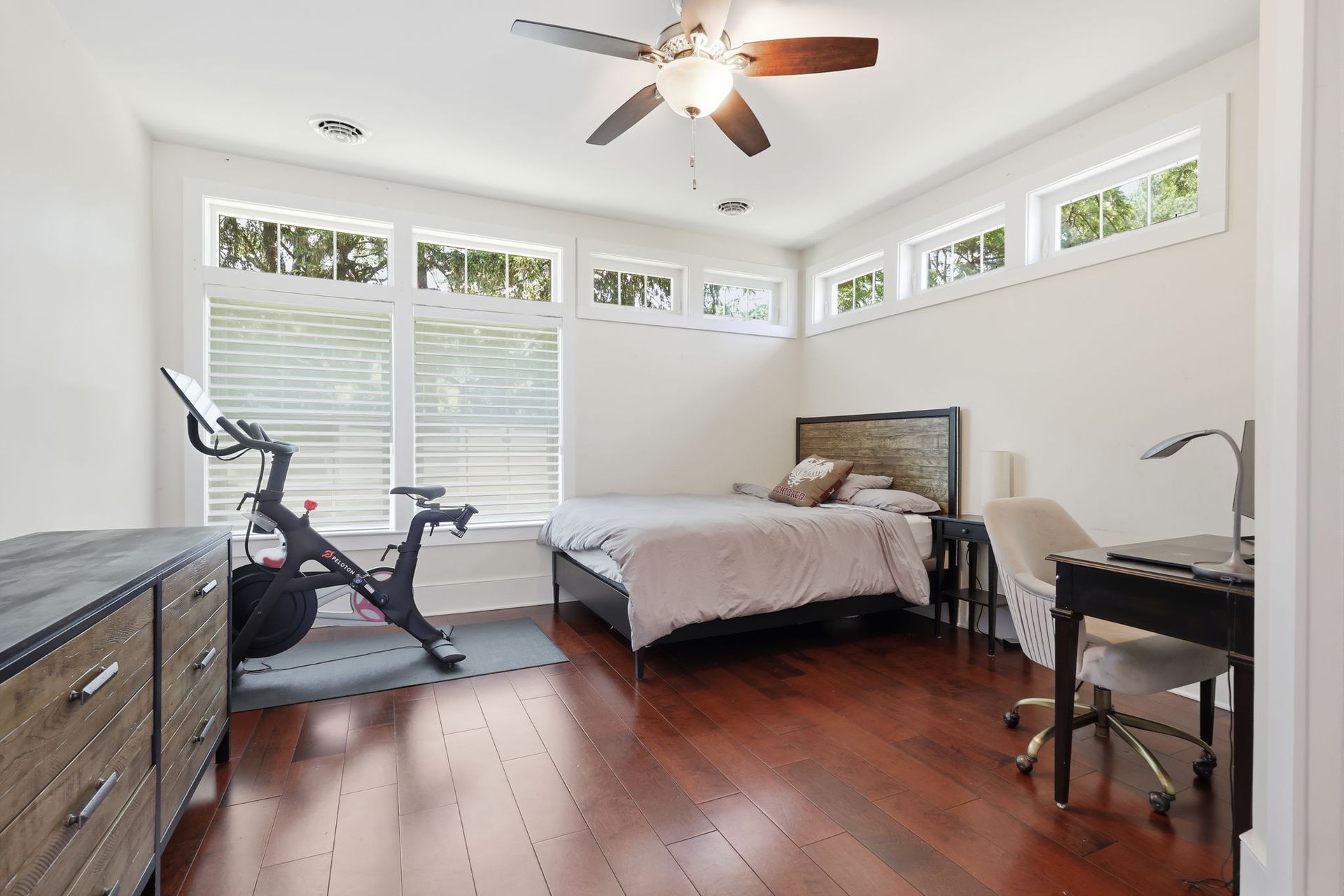 Bright bedroom with a bed, desk, exercise bike, and large windows with white blinds