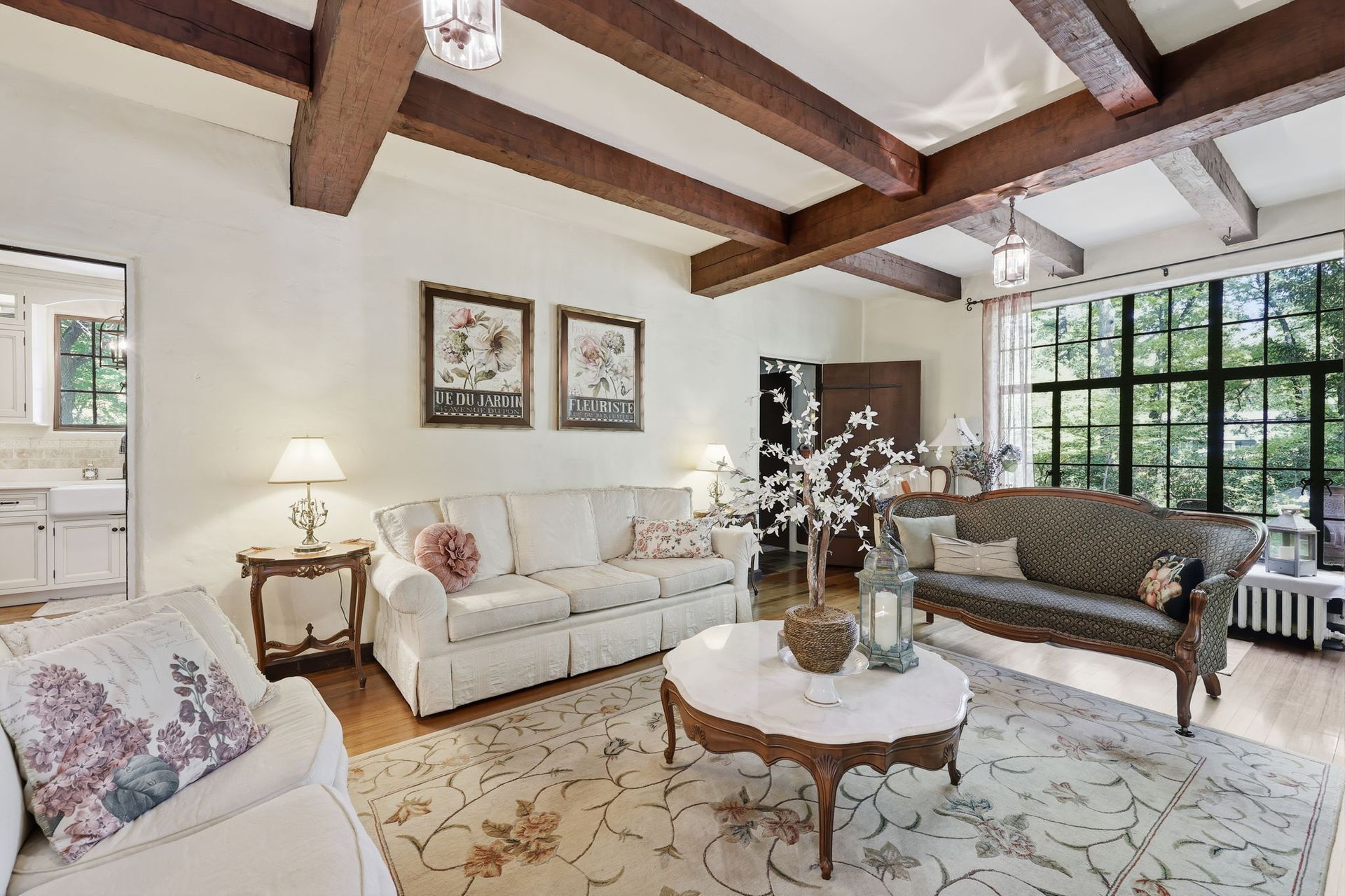 Bright living room with exposed wood beams, white sofa, patterned rug, and large windows overlooking greenery