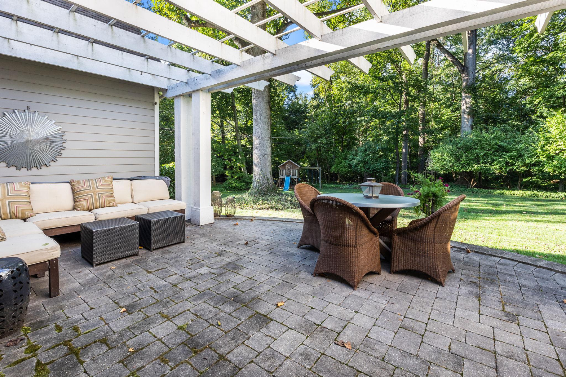 Outdoor patio with seating and table under a white pergola, set against a green lawn and trees.