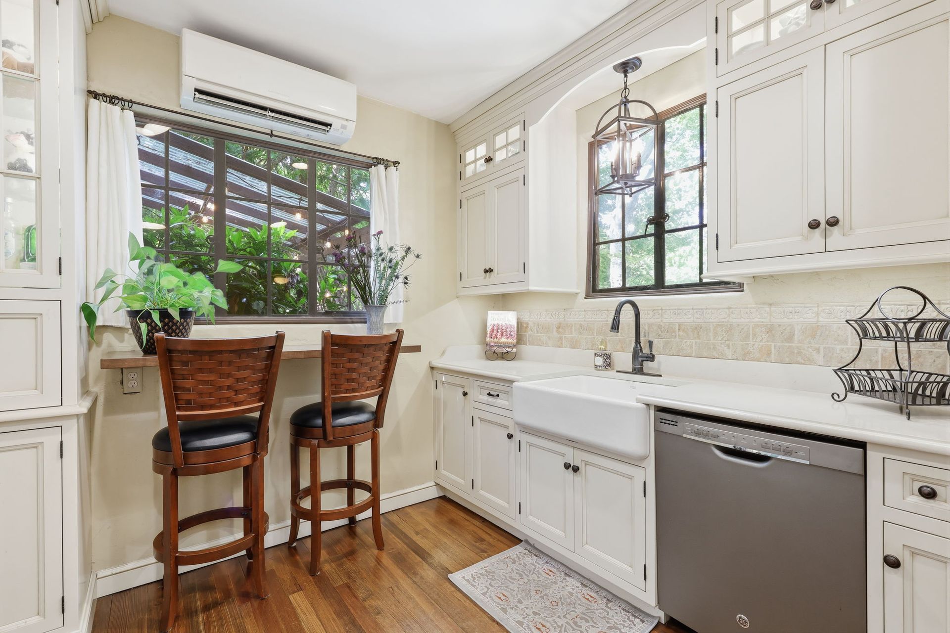 Bright kitchen with white cabinets, stainless dishwasher, wood chairs, and a window above the sink