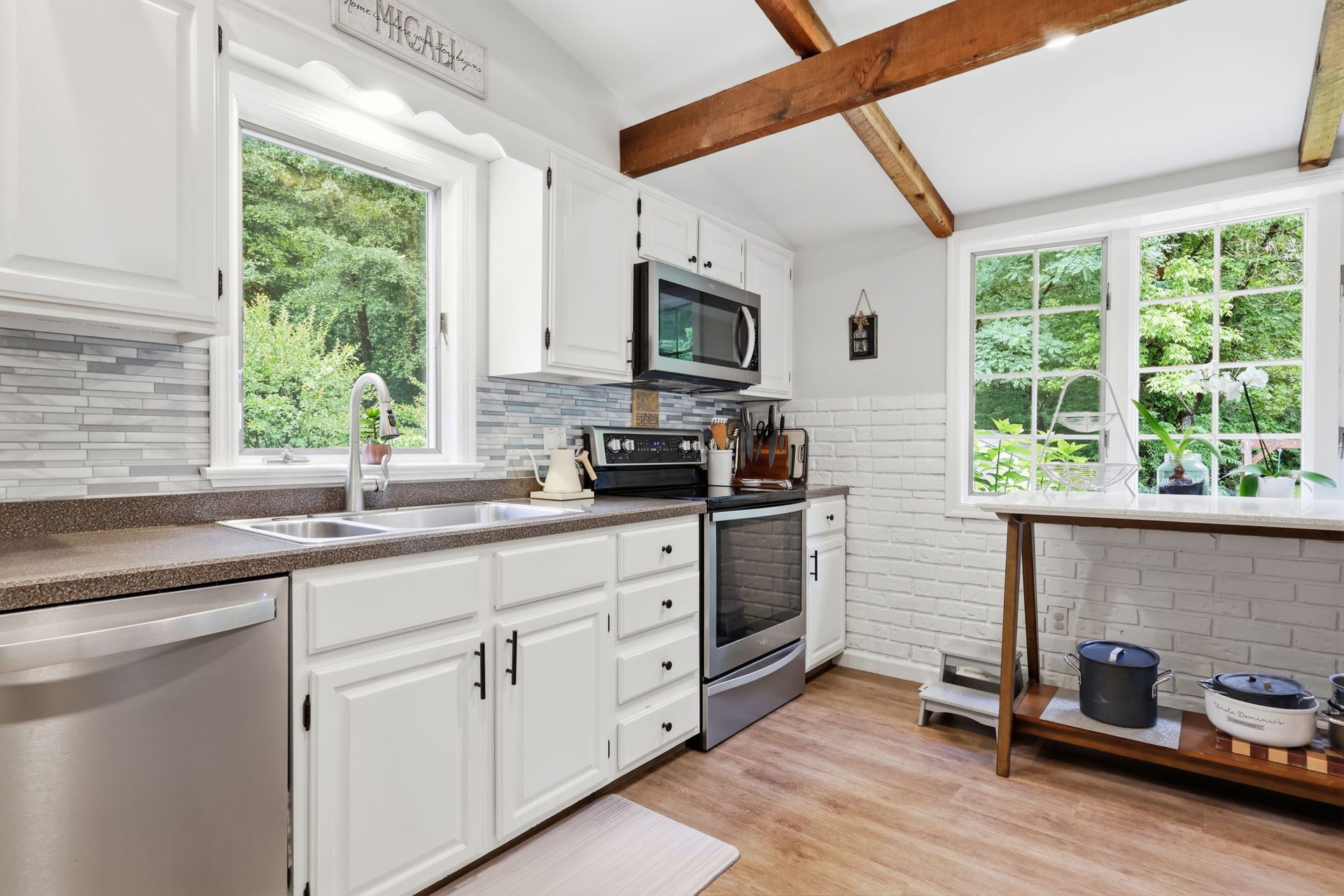 Bright white kitchen with wood beams, gray countertops, stainless appliances, and a window over the sink.