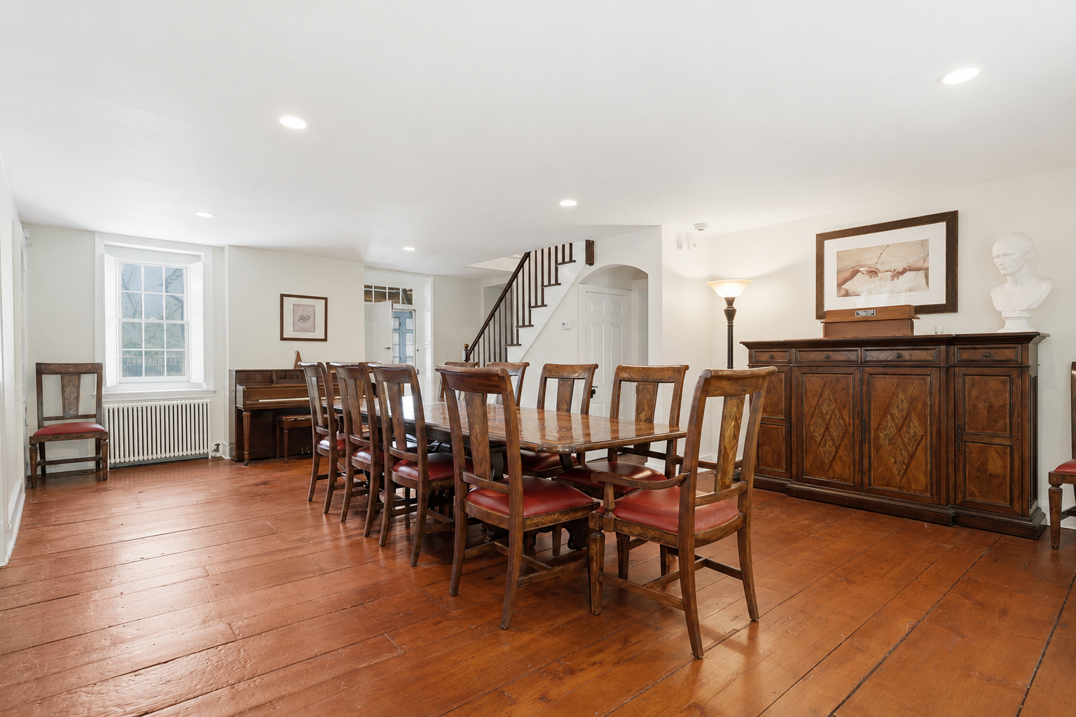Bright dining room with long wooden table, chairs, sideboard, staircase, and framed artwork.