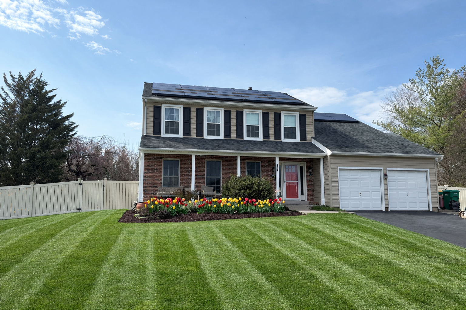A two-story house with solar panels, a manicured lawn, a colorful flower bed, and a two-car garage under a blue sky.