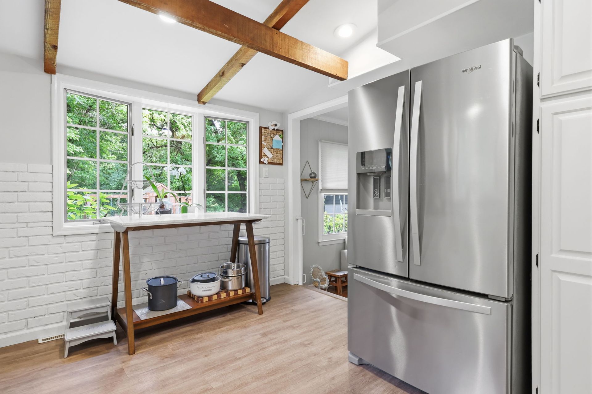 Bright kitchen with stainless steel fridge, white cabinets, exposed wood beam, and window over wooden cart