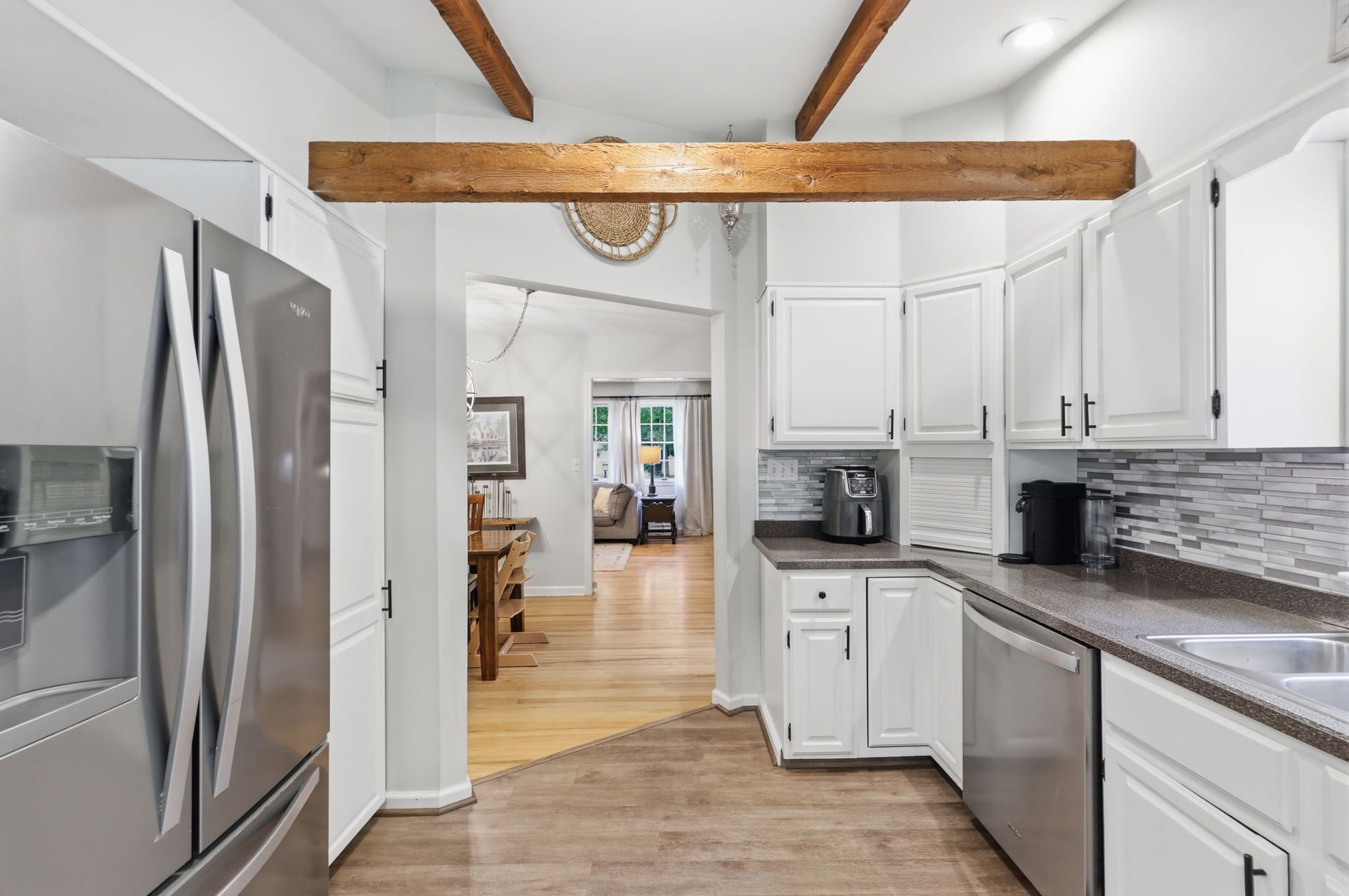 Modern white kitchen with stainless steel appliances, exposed wood beams, and a view into a dining room