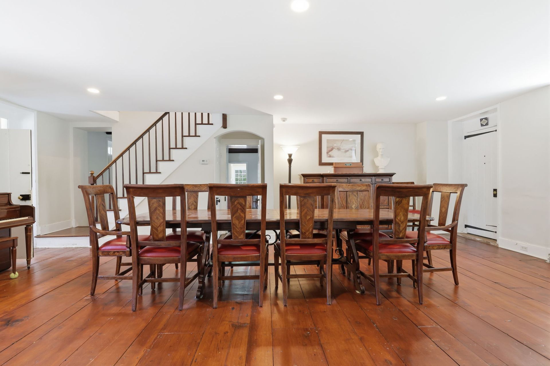 Dining room with a long wooden table and chairs on a polished hardwood floor, white walls, and staircase in back