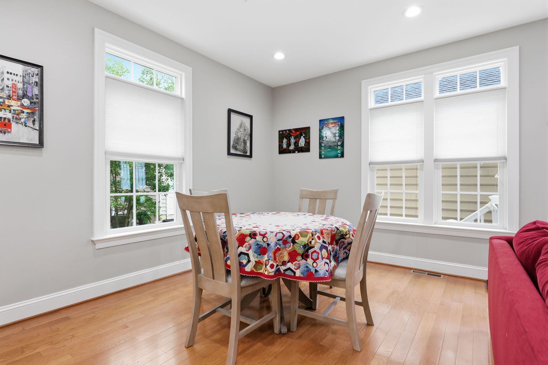 Bright dining room with wooden table, colorful cloth, white windows, and red sofa by hardwood floor