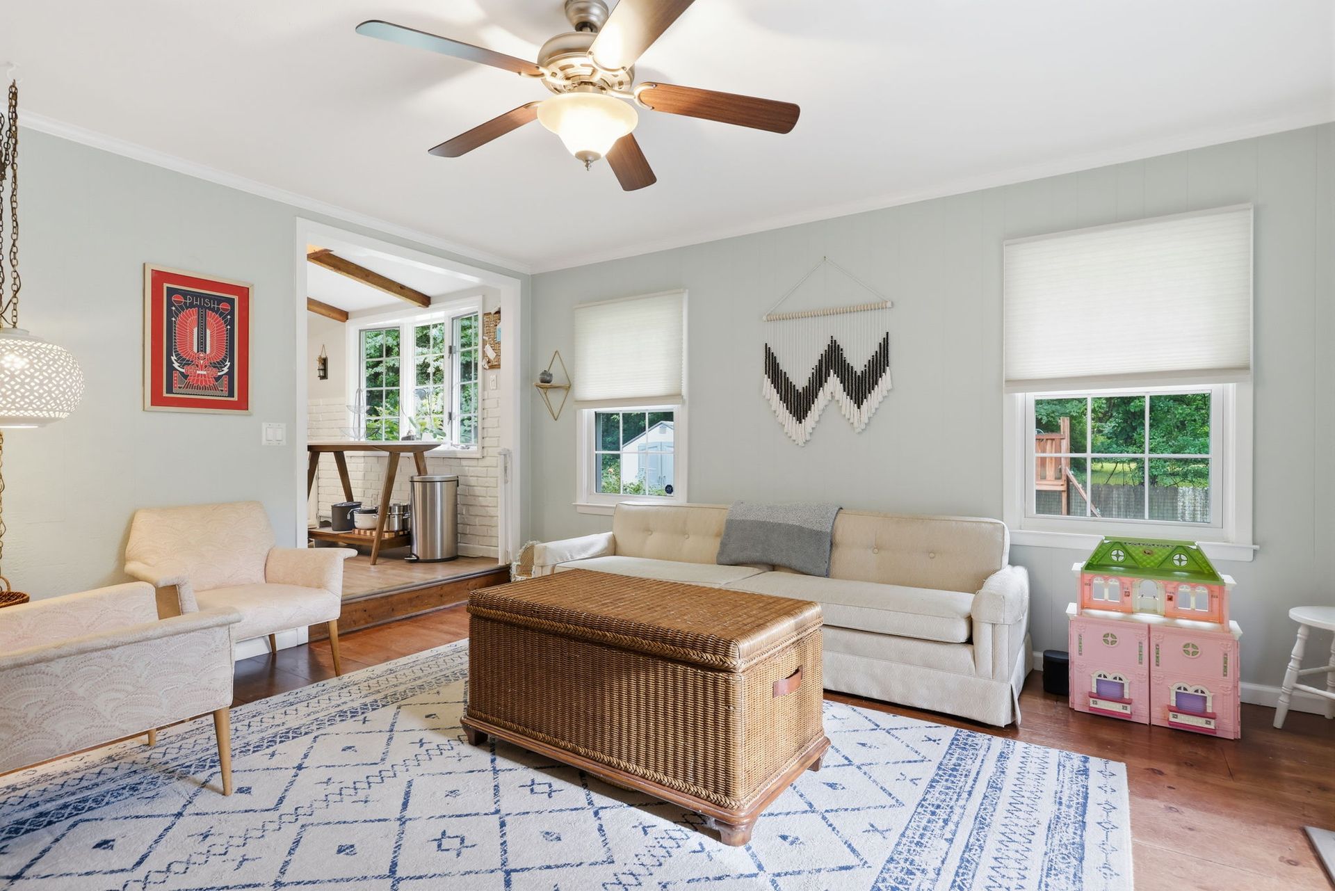 Bright living room with white sofas, wicker coffee table, patterned rug, and ceiling fan by windows.