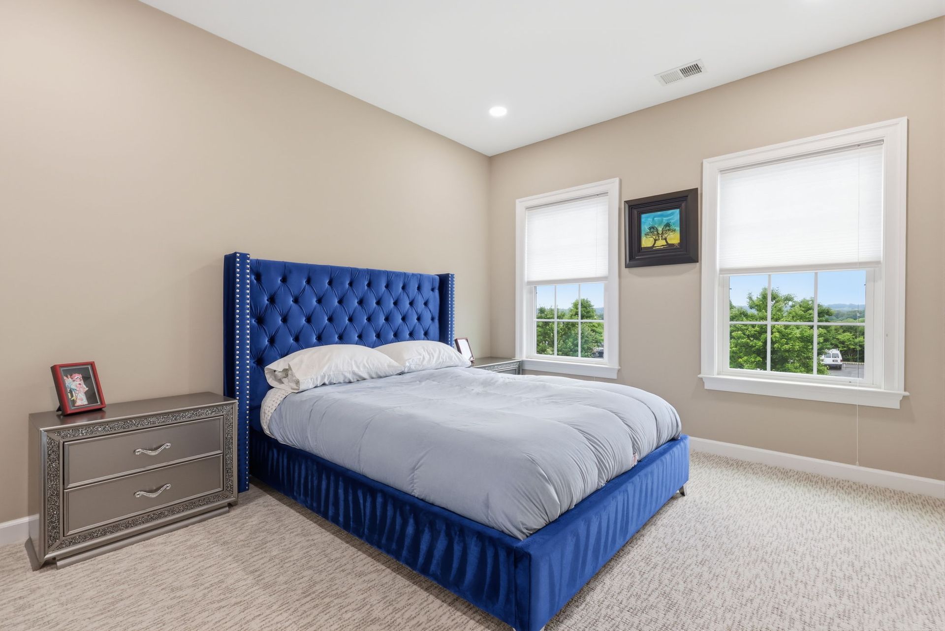Blue upholstered bed in a bright beige bedroom with two windows and a gray nightstand