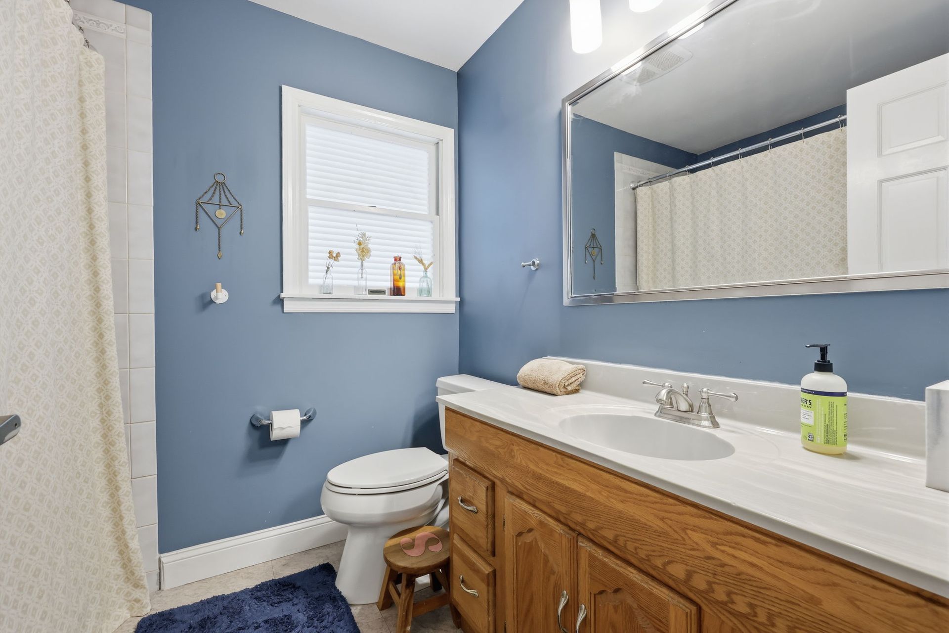 Small blue bathroom with white vanity, toilet, window, and mirror above a wood cabinet.