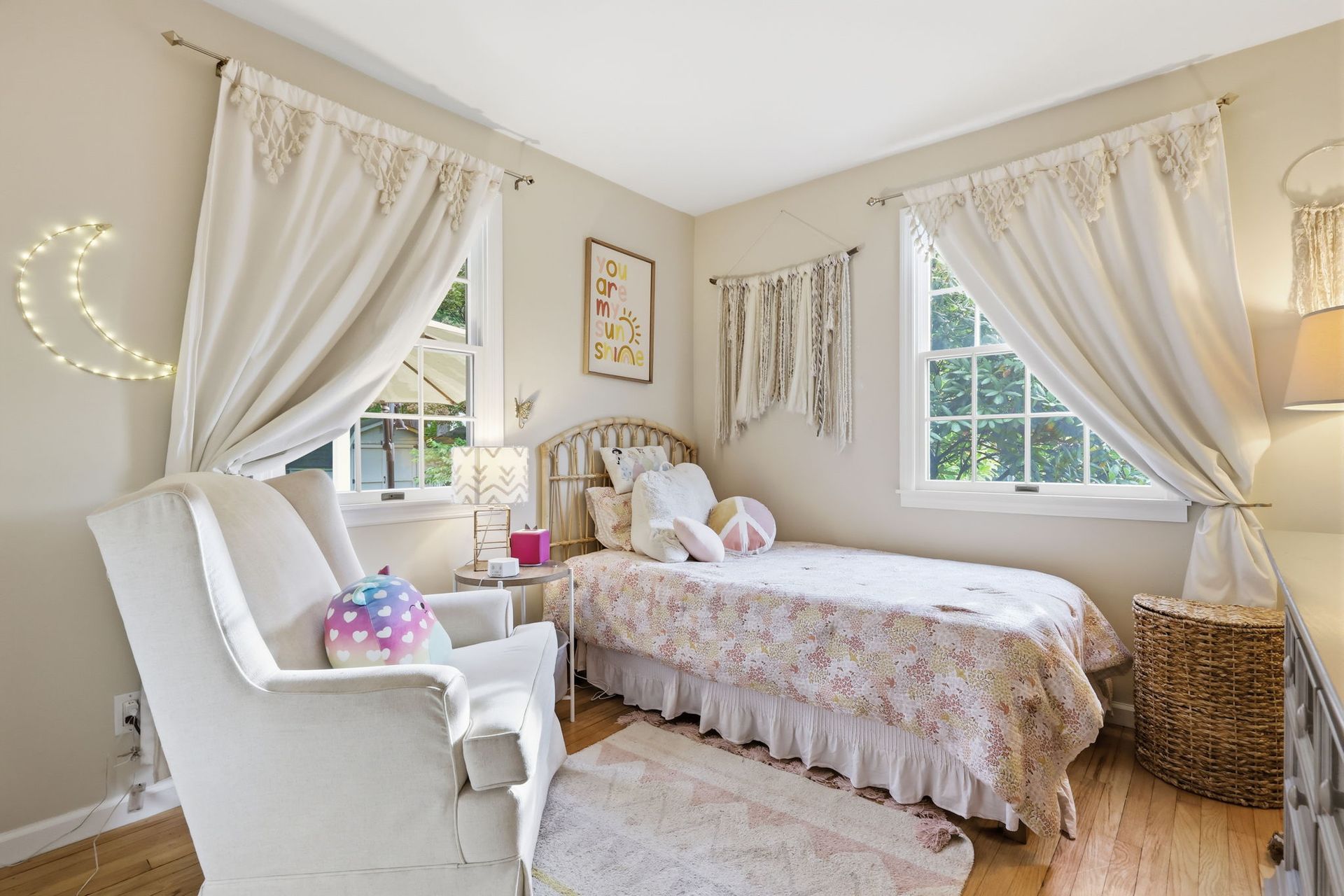 Bright pastel bedroom with a twin bed, white chair, draped windows, and soft fairy lights