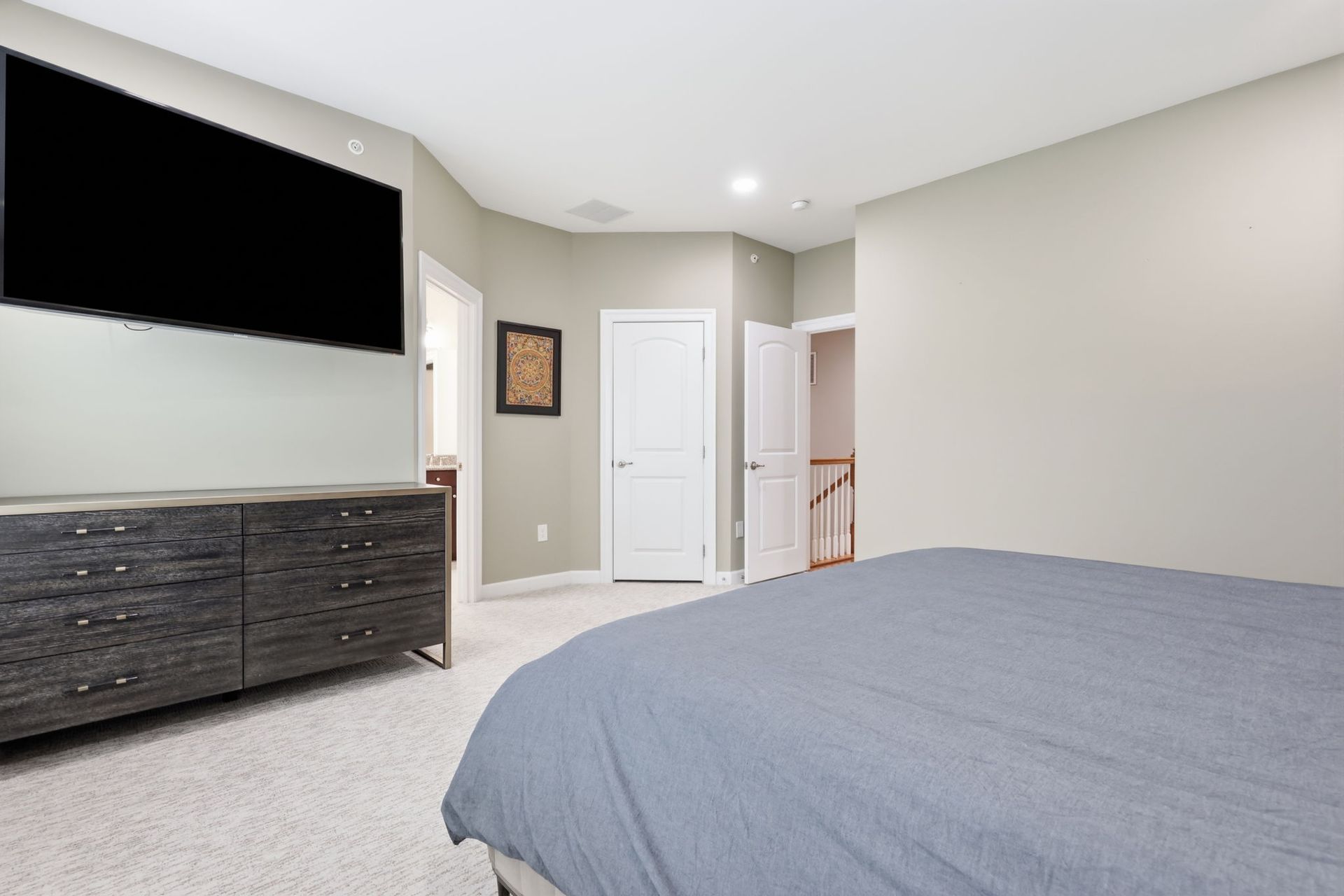 Bedroom with a bed, dresser, wall-mounted TV, and white door in a neutral-toned interior.