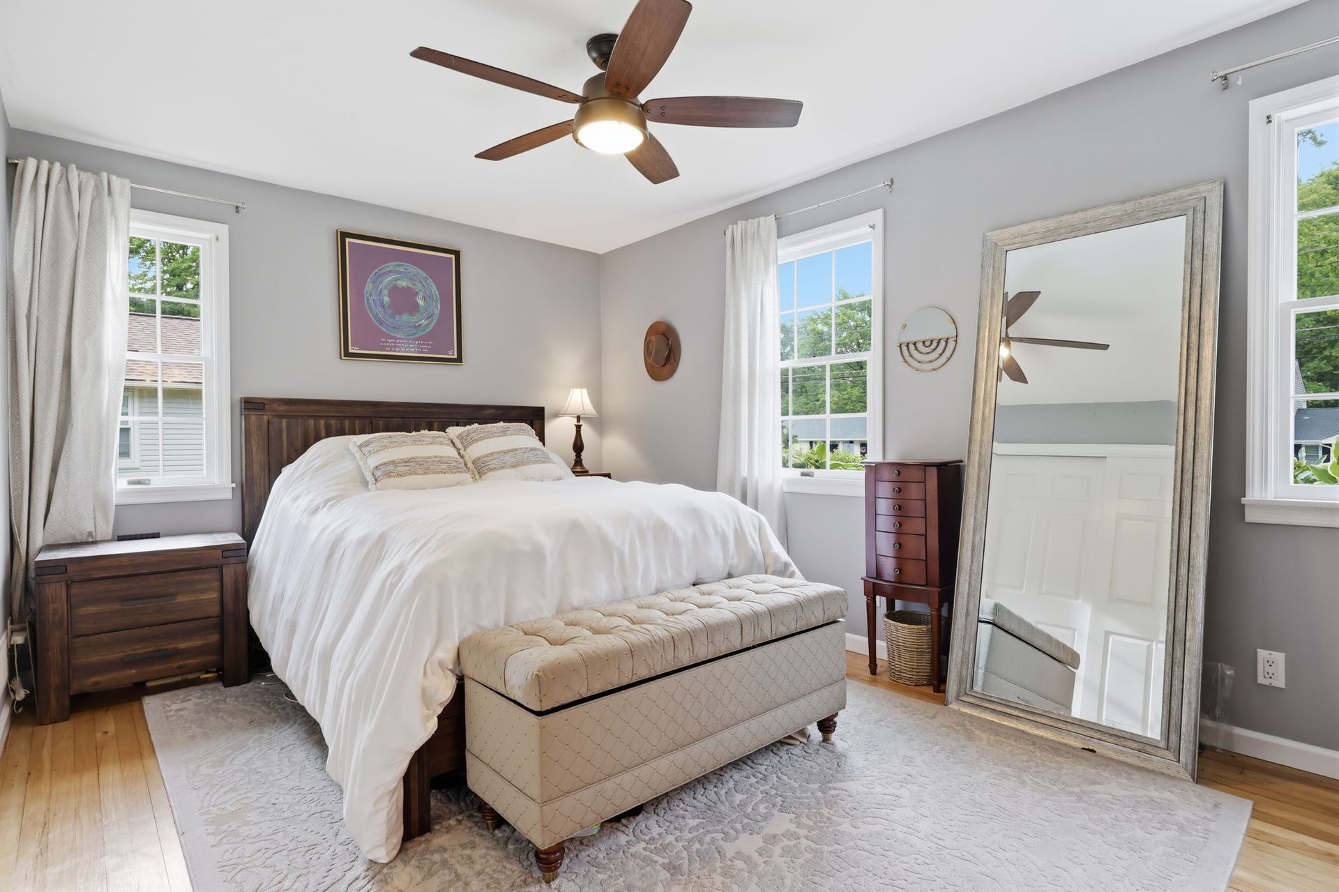 Bright bedroom with white bed, wooden nightstands, bench, mirror, and ceiling fan in a light gray room