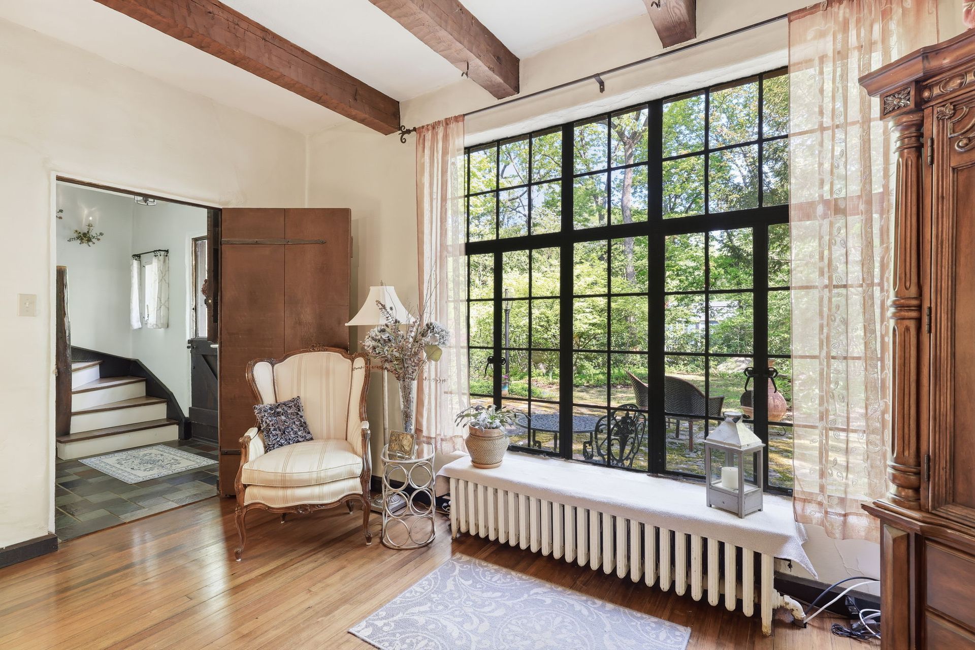 Bright living room with large window, wicker chair, radiator, and wooden beams.