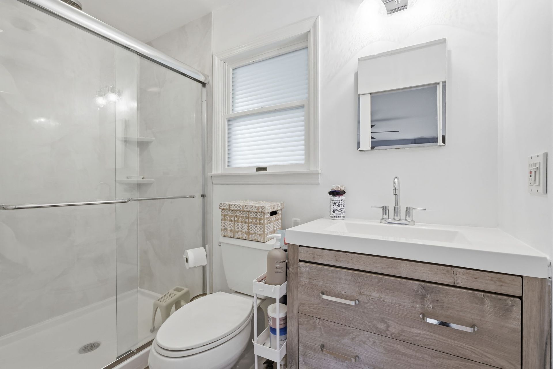 Bright white bathroom with glass shower, toilet, window, and wood vanity with sink and mirror.