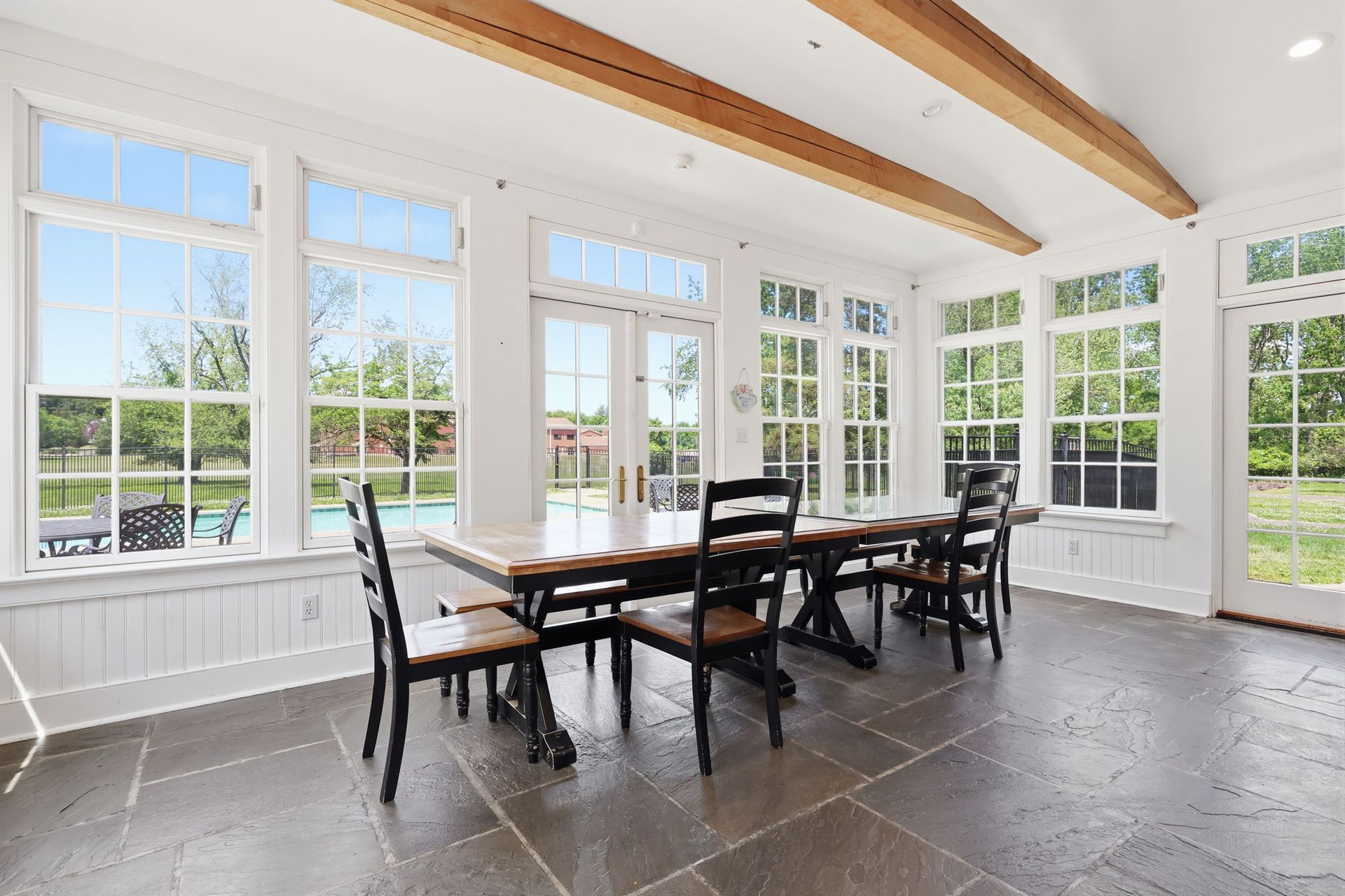 Bright sunroom dining area with a table, chairs, white walls, and large windows overlooking greenery