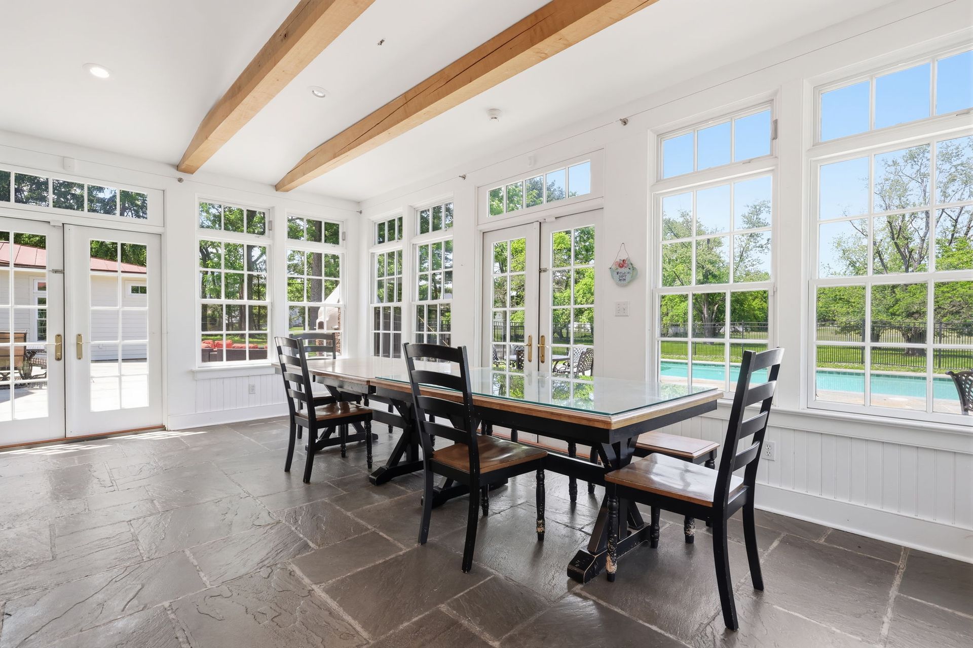 Bright dining room with large windows, wooden beams, and a table set with chairs overlooking a pool.