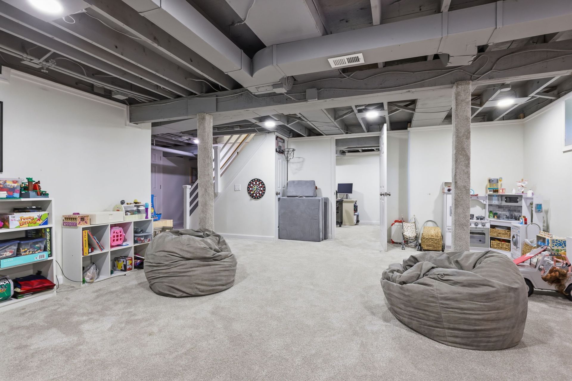 Bright basement playroom with gray bean bags, toys, shelves, and exposed ceiling beams