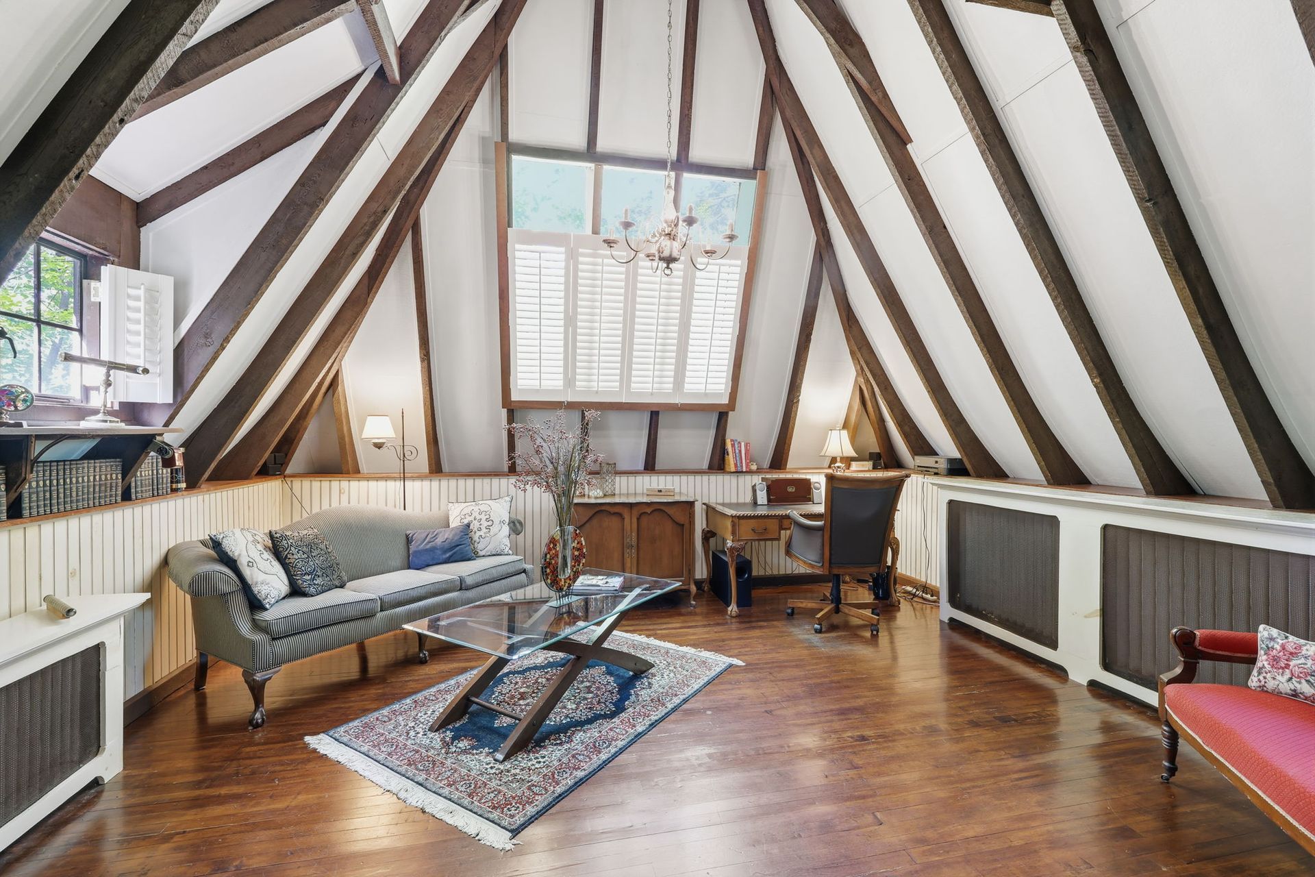 A cozy attic living room with exposed beams, a sofa, chairs, rug, and large central window.