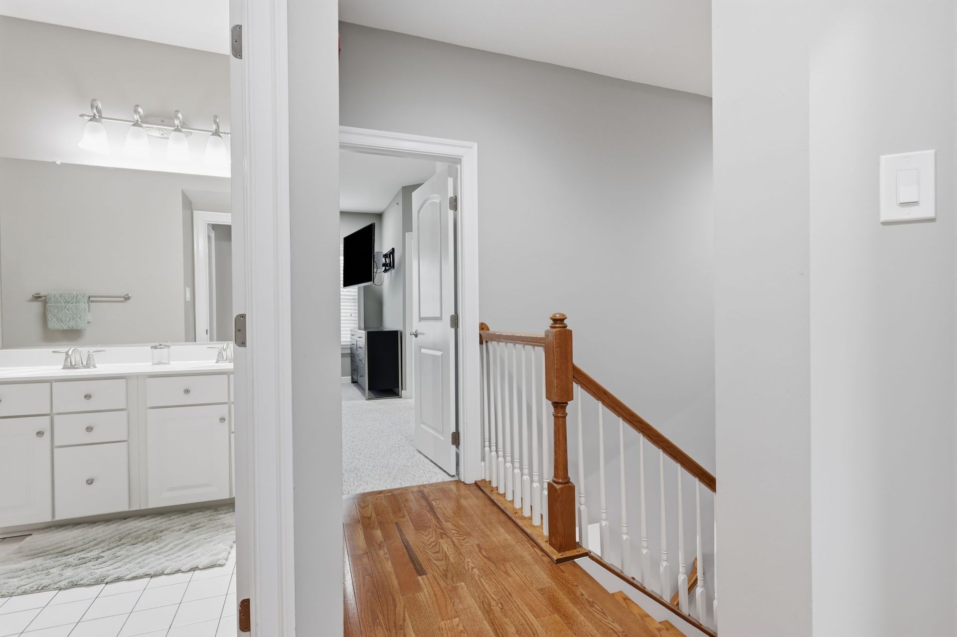 Bright hallway with hardwood floor, white railing, and a bathroom vanity visible through an open doorway.