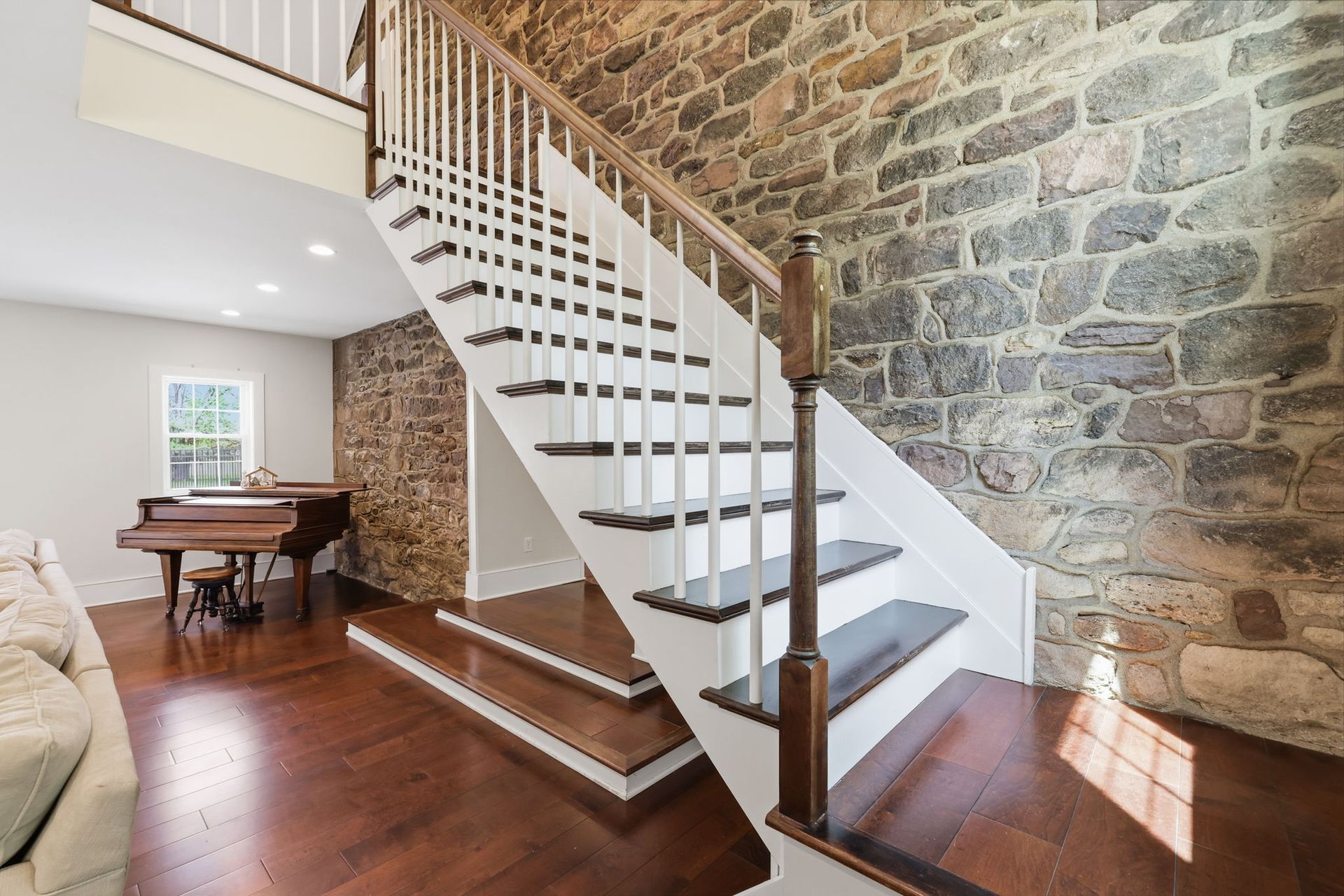Stone staircase with white railings in a sunlit room with hardwood floors and a piano.