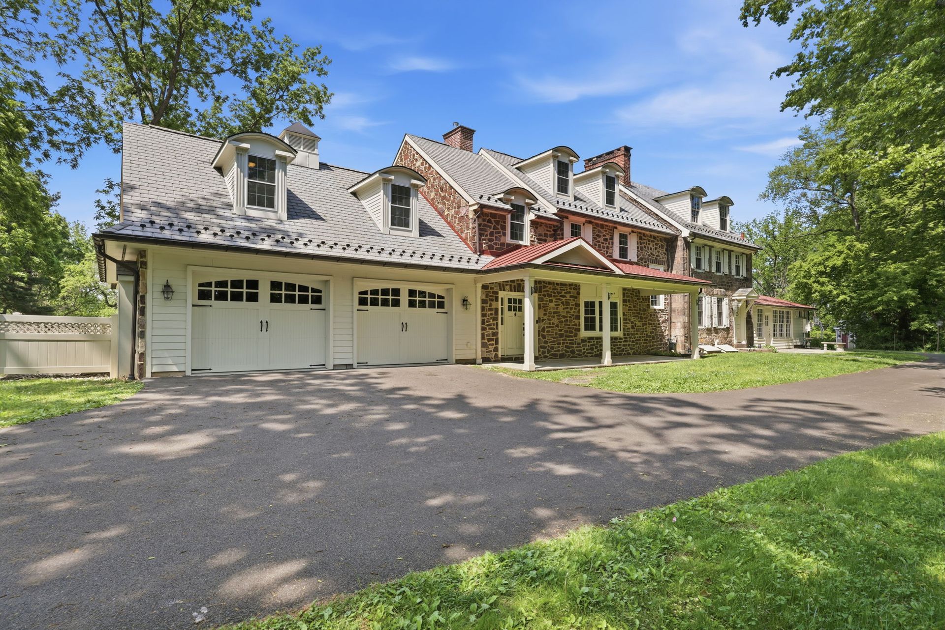 Large stone house with garage doors, dormers, and tree-lined front drive under a blue sky