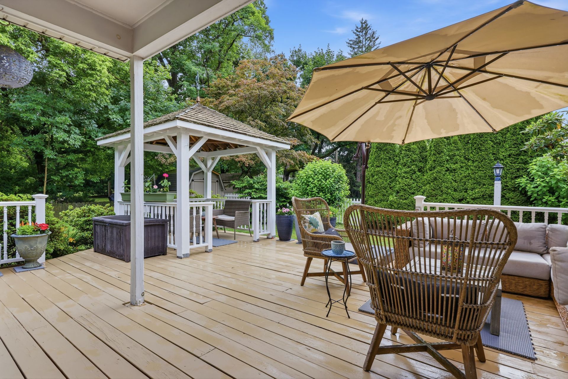 Covered patio with wicker seating, beige umbrella, and a backyard gazebo surrounded by greenery