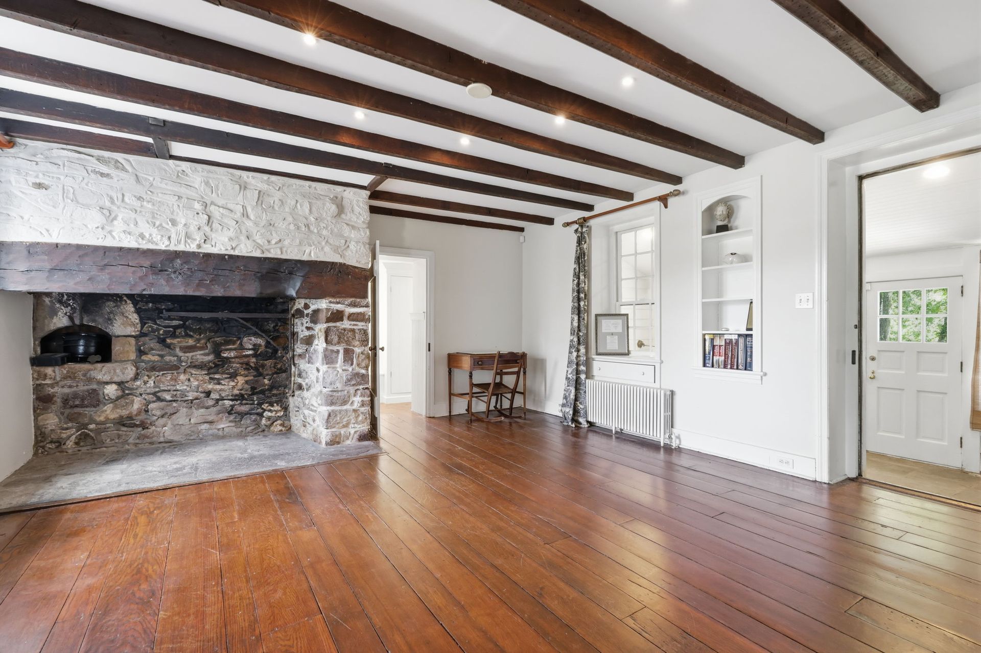 Empty rustic room with exposed ceiling beams, brick fireplace, wooden floor, and white walls