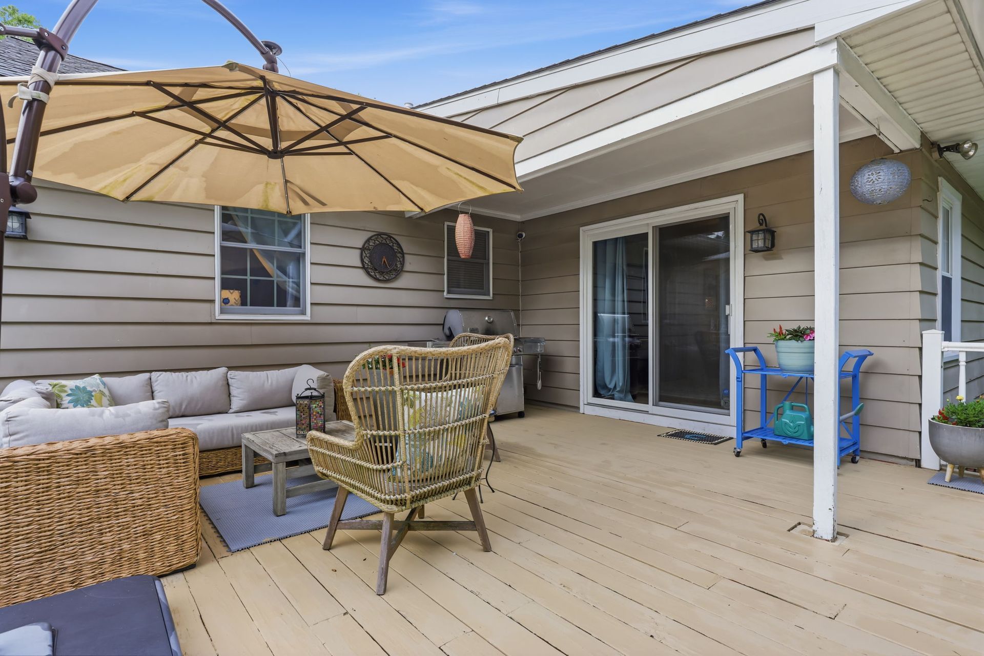 Covered patio with wicker seating, large beige umbrella, and sliding glass doors beside a beige house.