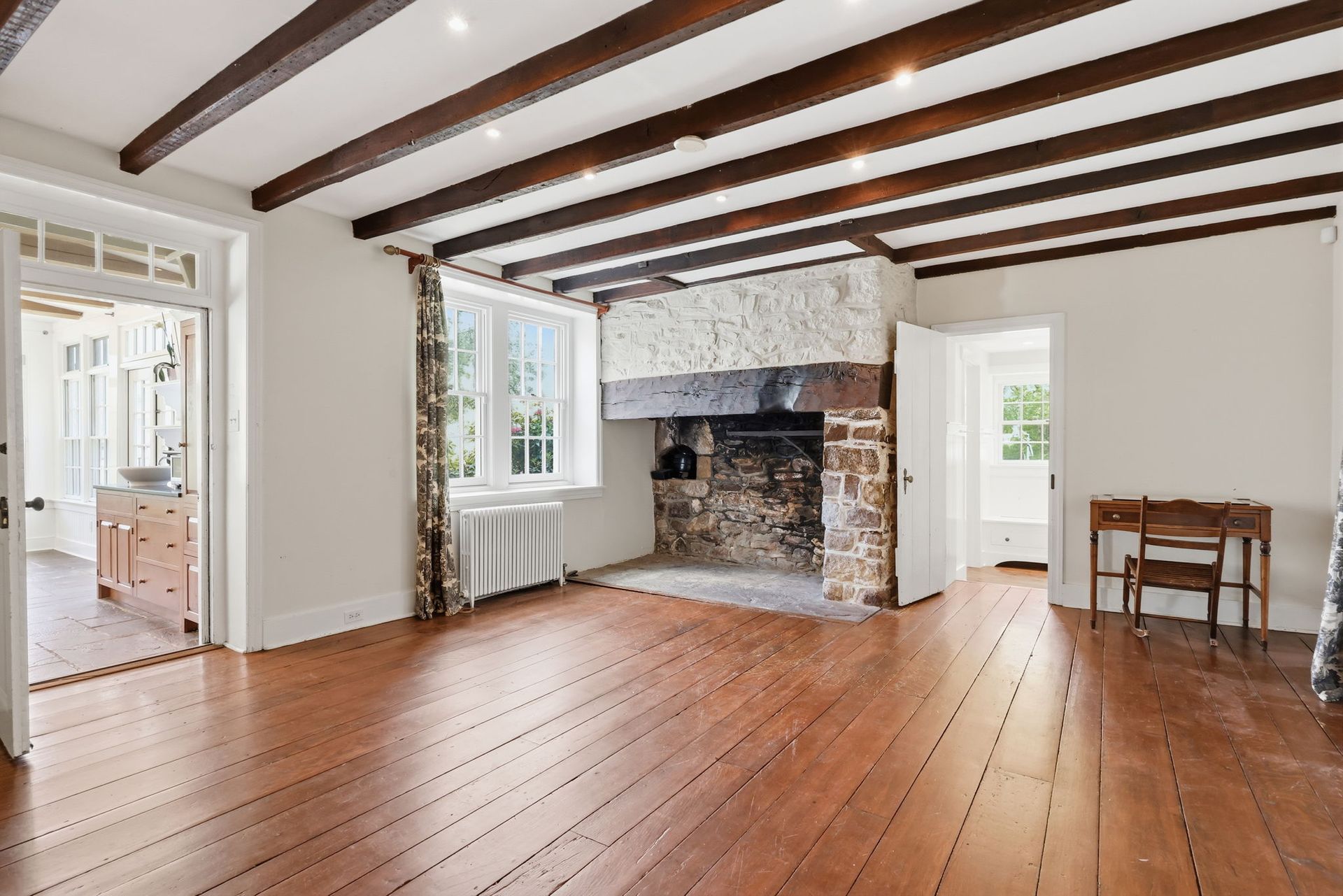 Empty room with wooden floors, exposed beams, stone fireplace, and bright windows
