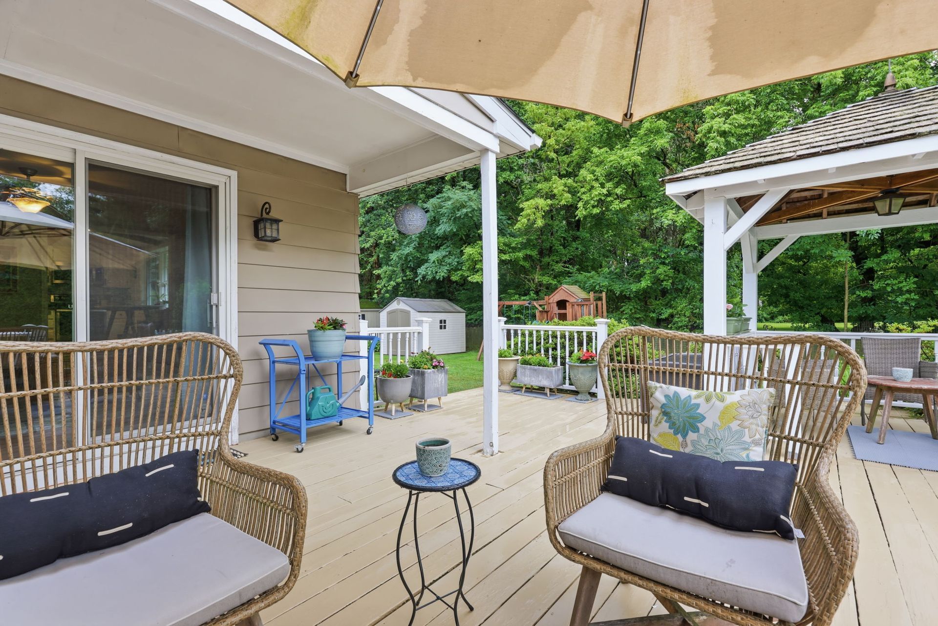Patio with wicker chairs, small tables, umbrella, and grill beside a house, overlooking a green yard