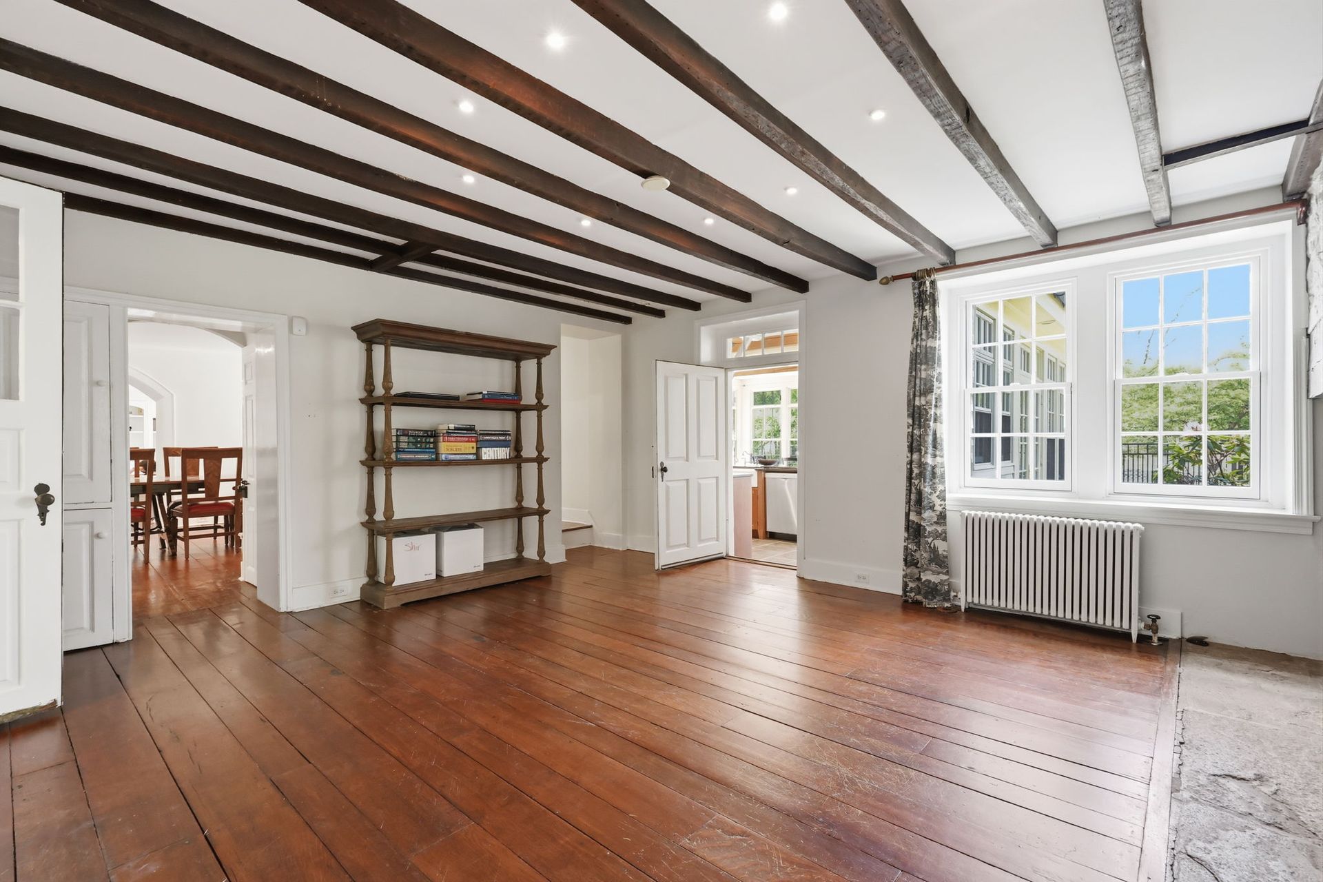 Bright empty room with wooden floors, white walls, exposed ceiling beams, shelves, and large windows.