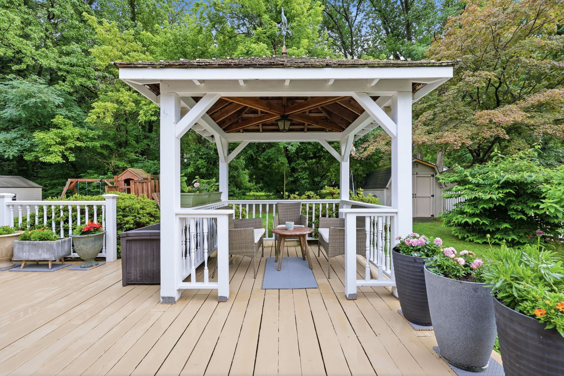 White garden gazebo on a wooden deck, surrounded by potted flowers and green trees